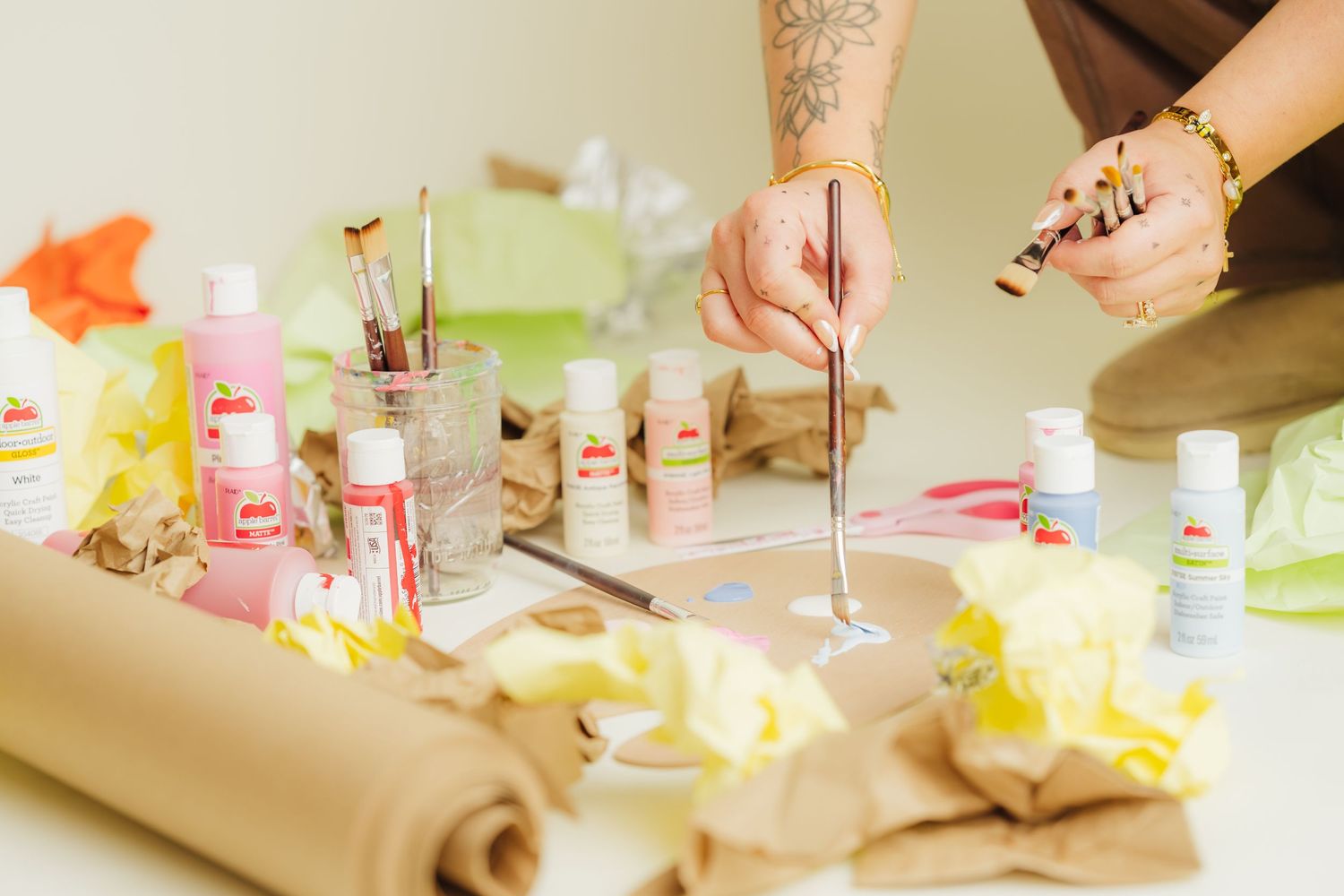 Artist painting at a craft table with pink acrylic paints, brushes, and crumpled paper in a bright studio.