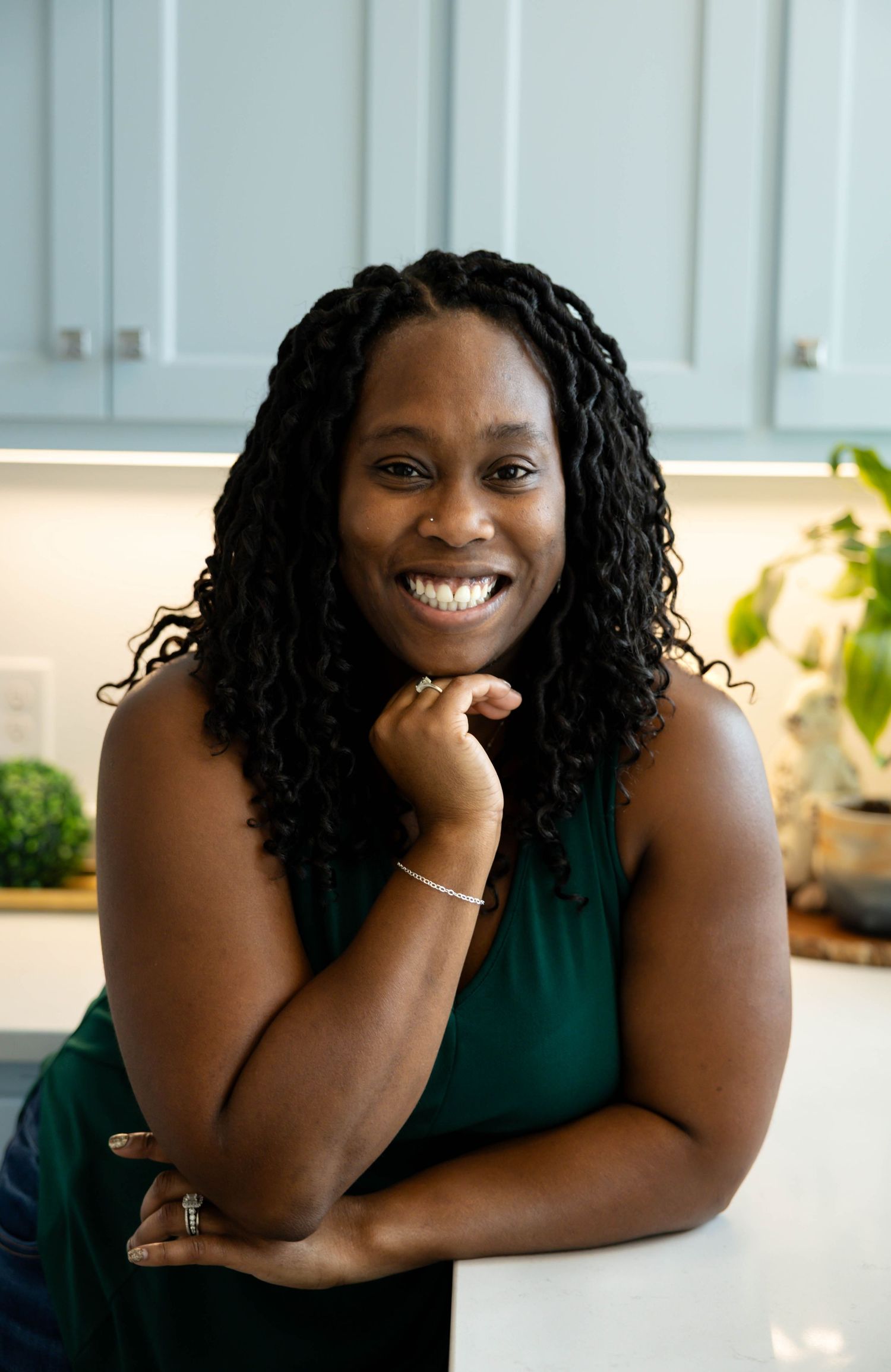 A person in a dark green sleeveless top smiles warmly while sitting at a white table in a bright indoor setting.