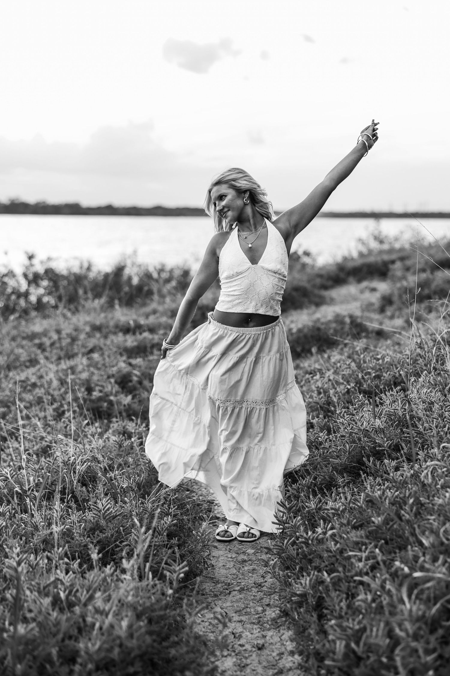 Black and white photo of a joyful blonde woman in white maxi skirt dancing on a nature path by the water.