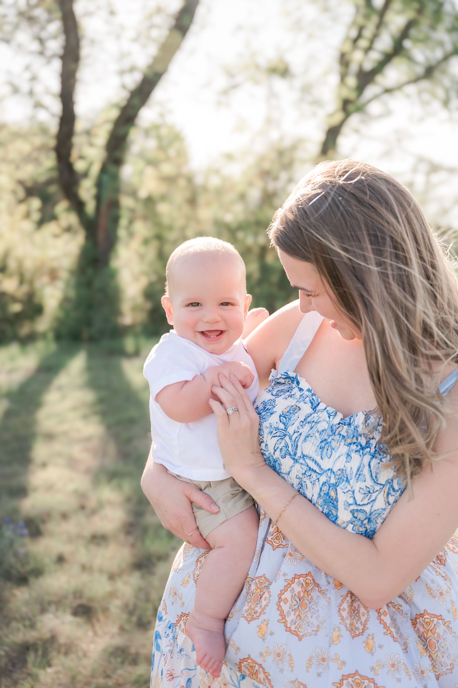 A smiling baby in white shirt held by a woman in floral dress outdoors in golden sunlight. Northlake Tx, Knot & Wing Photography, Argyle, Tx