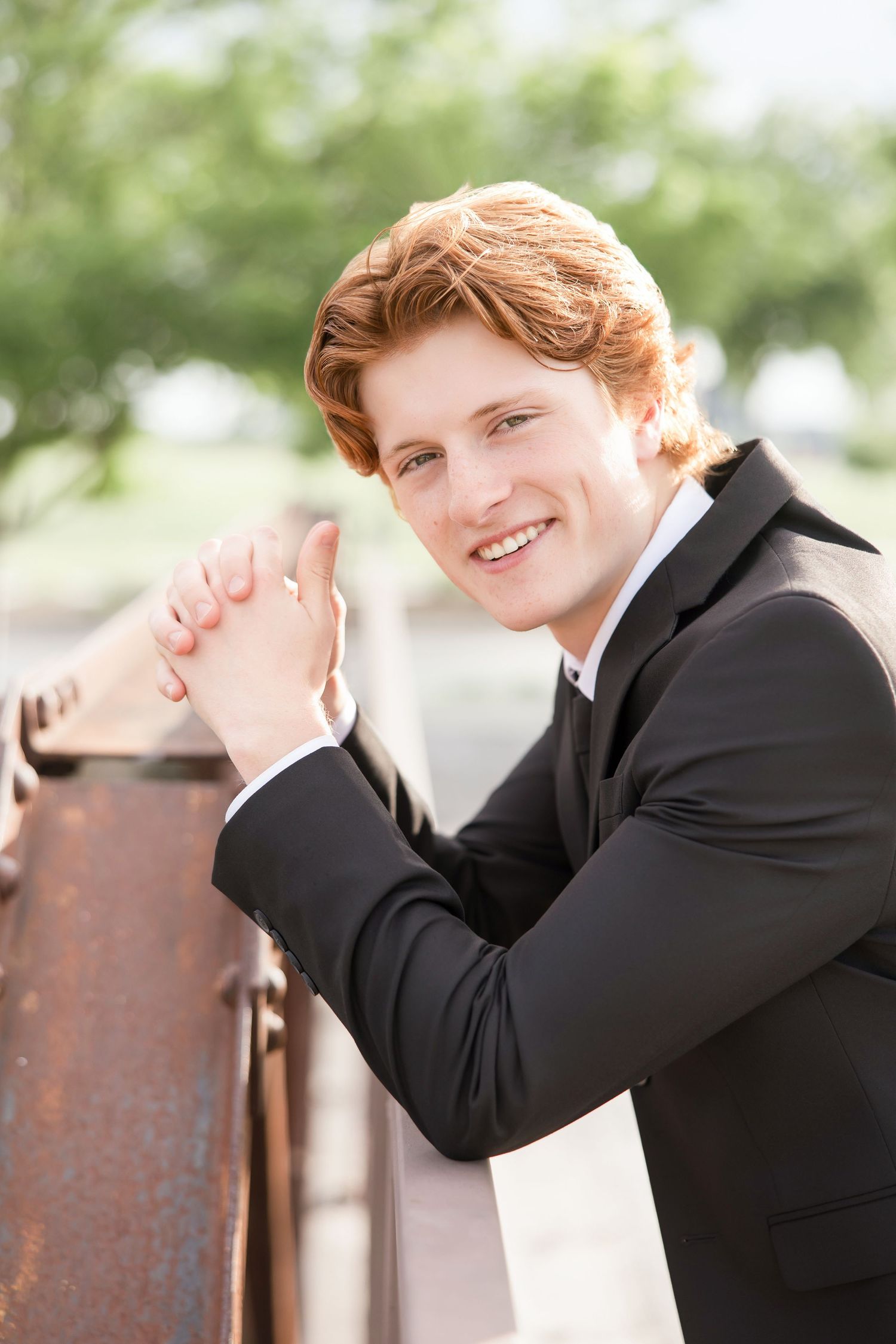 Seniors 2026 Photography .Smiling young man with red hair in a black suit leaning on a rustic railing outdoors.  Knot & Wing Photography