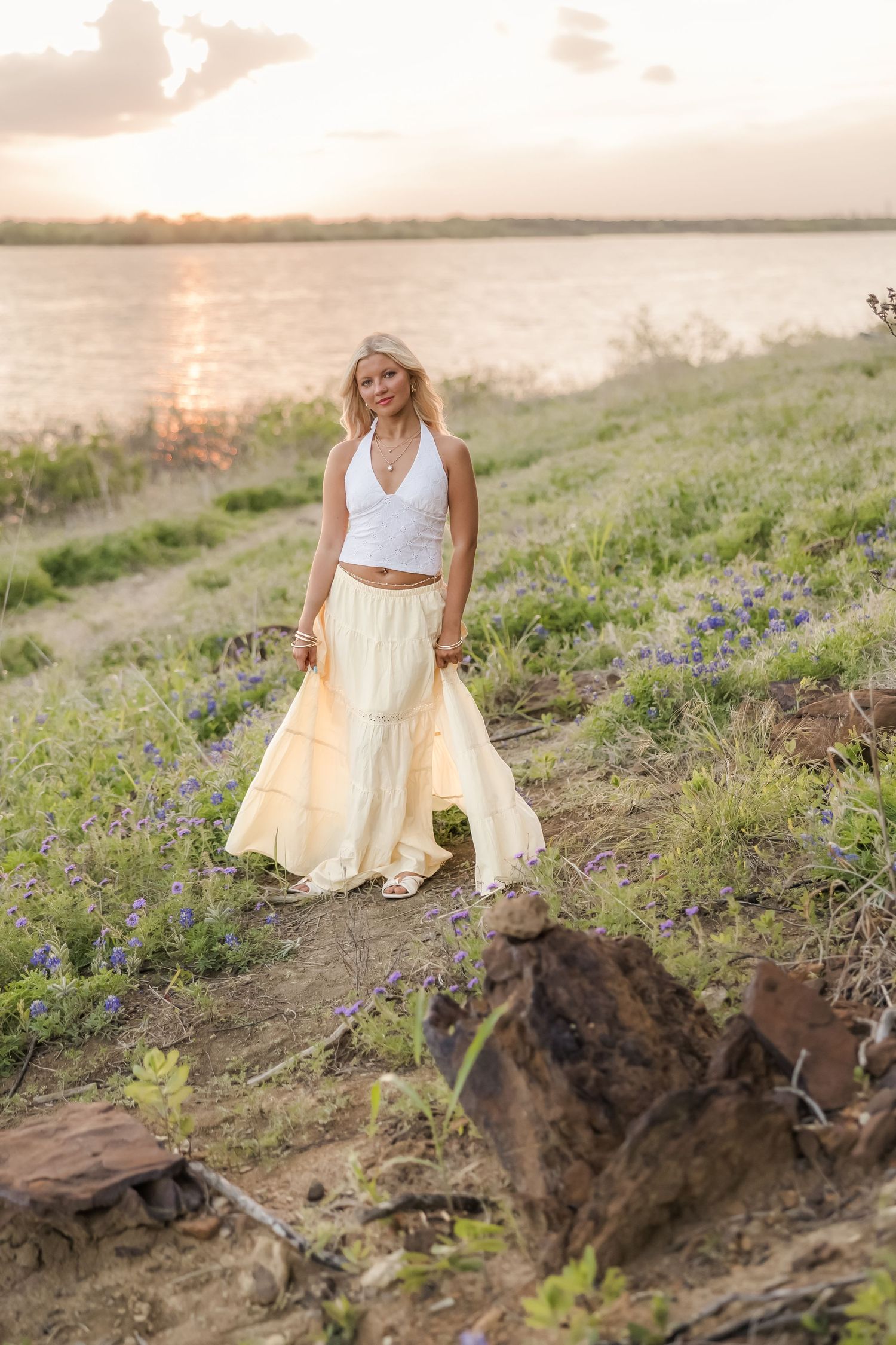 Blonde woman in white crop top and yellow maxi skirt standing near a river at sunset with wildflowers.