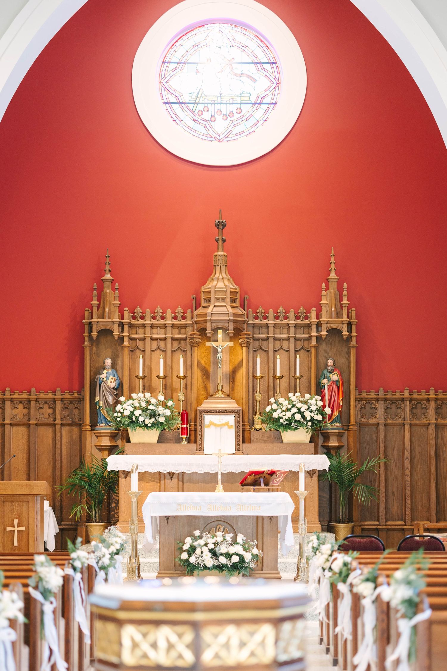 Ornate wooden church altar with statues, candles, and white floral arrangements beneath a red wall and round stained glass window.
