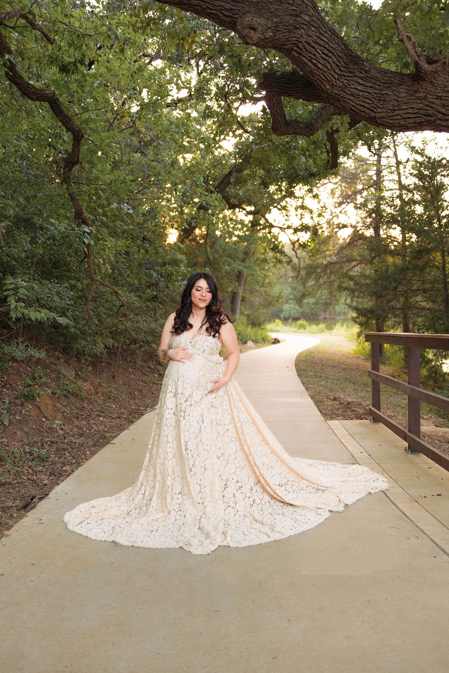 Pregnant woman in white lace gown poses on a tree-lined path at golden hour in a serene outdoor setting.