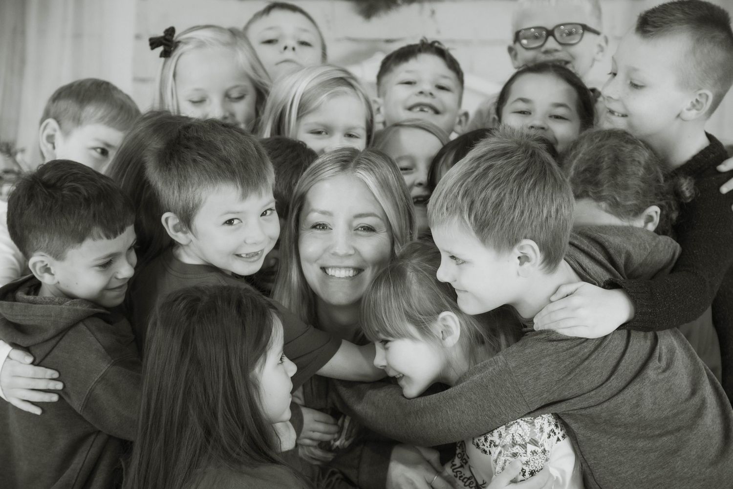 Black and white photo of a smiling woman surrounded by a large group of happy children hugging her.