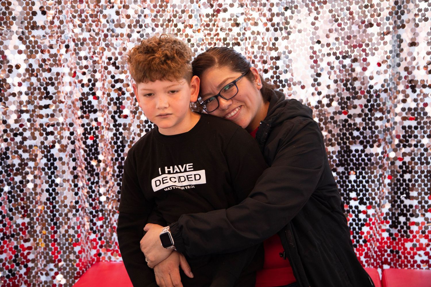 A boy in a black graphic tee and a woman with glasses pose together in front of a shiny sequin backdrop.