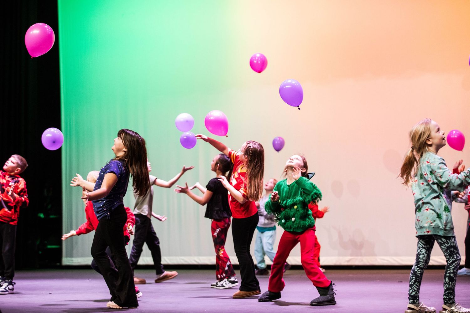 Children dancing on stage with colorful balloons during a festive holiday performance under green and pink lights.