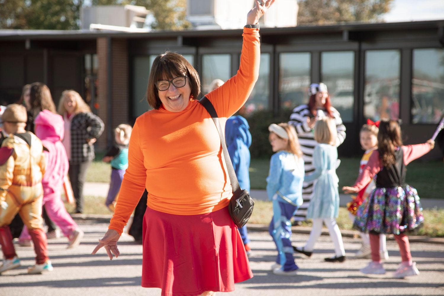 Person in orange Velma costume with raised arm at outdoor Halloween event with children in costumes.