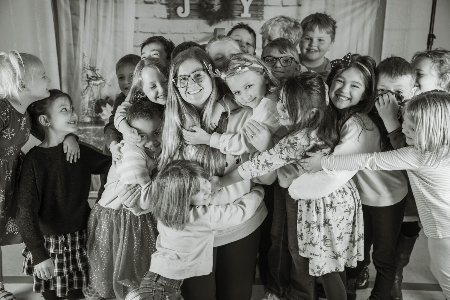 Black and white photo of a smiling woman surrounded by a large group of children hugging her indoors.