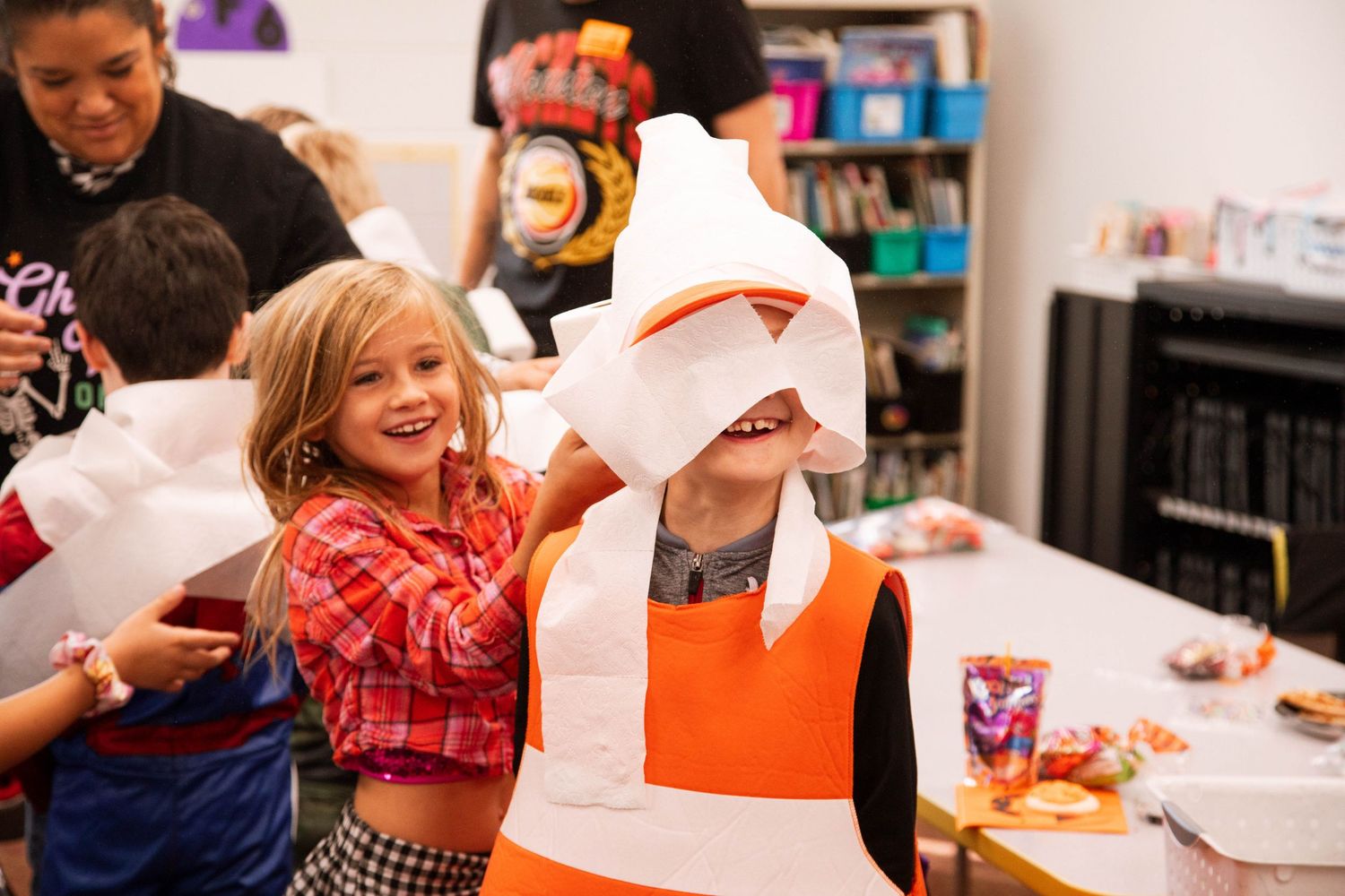 Children laughing and playing dress-up in colorful Halloween costumes during a festive classroom party.