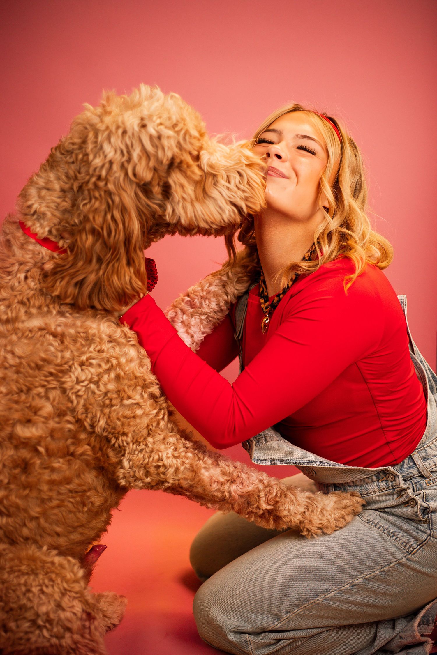 Young woman in red top and denim overalls sitting on pink background while golden doodle dog licks her cheek.