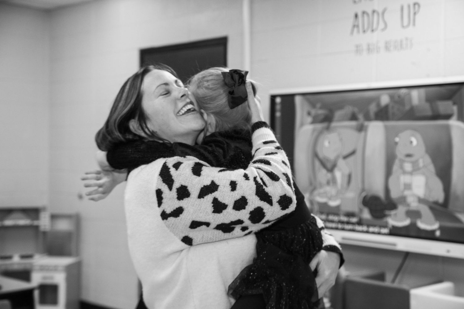 A woman in a cow-print sweater sharing a warm hug with a child in a classroom.