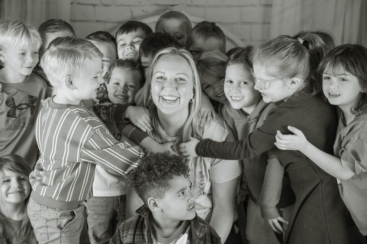 A smiling woman surrounded by hugging children in a black and white classroom photo.
