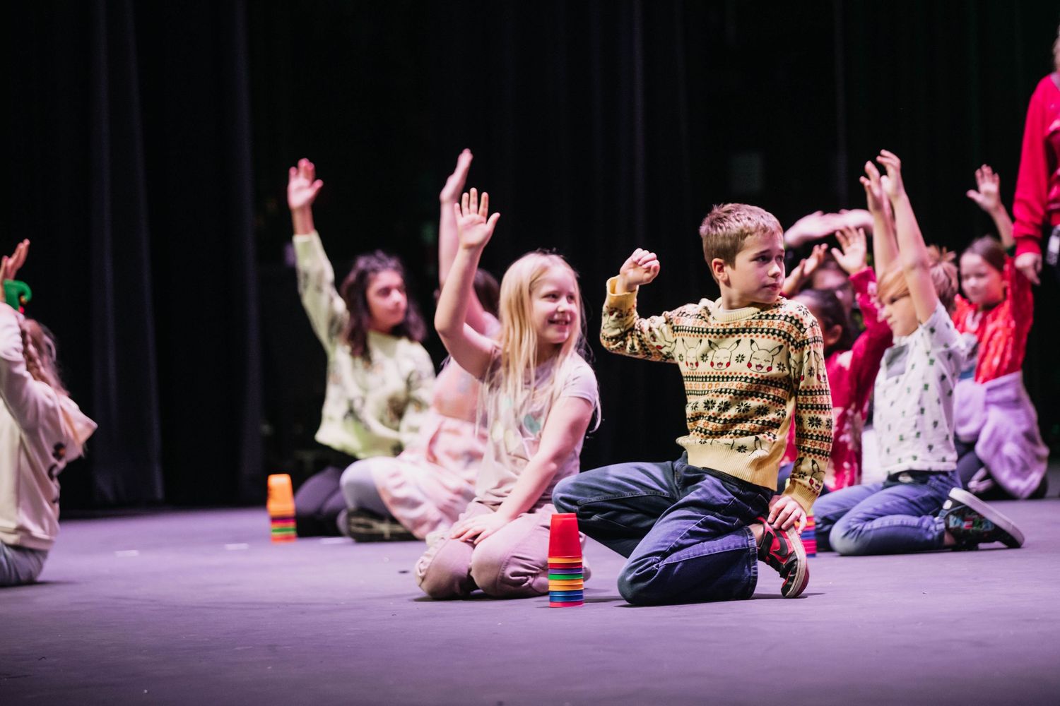 Children sitting on stage with hands raised during a fun kids theater performance under bright stage lights.