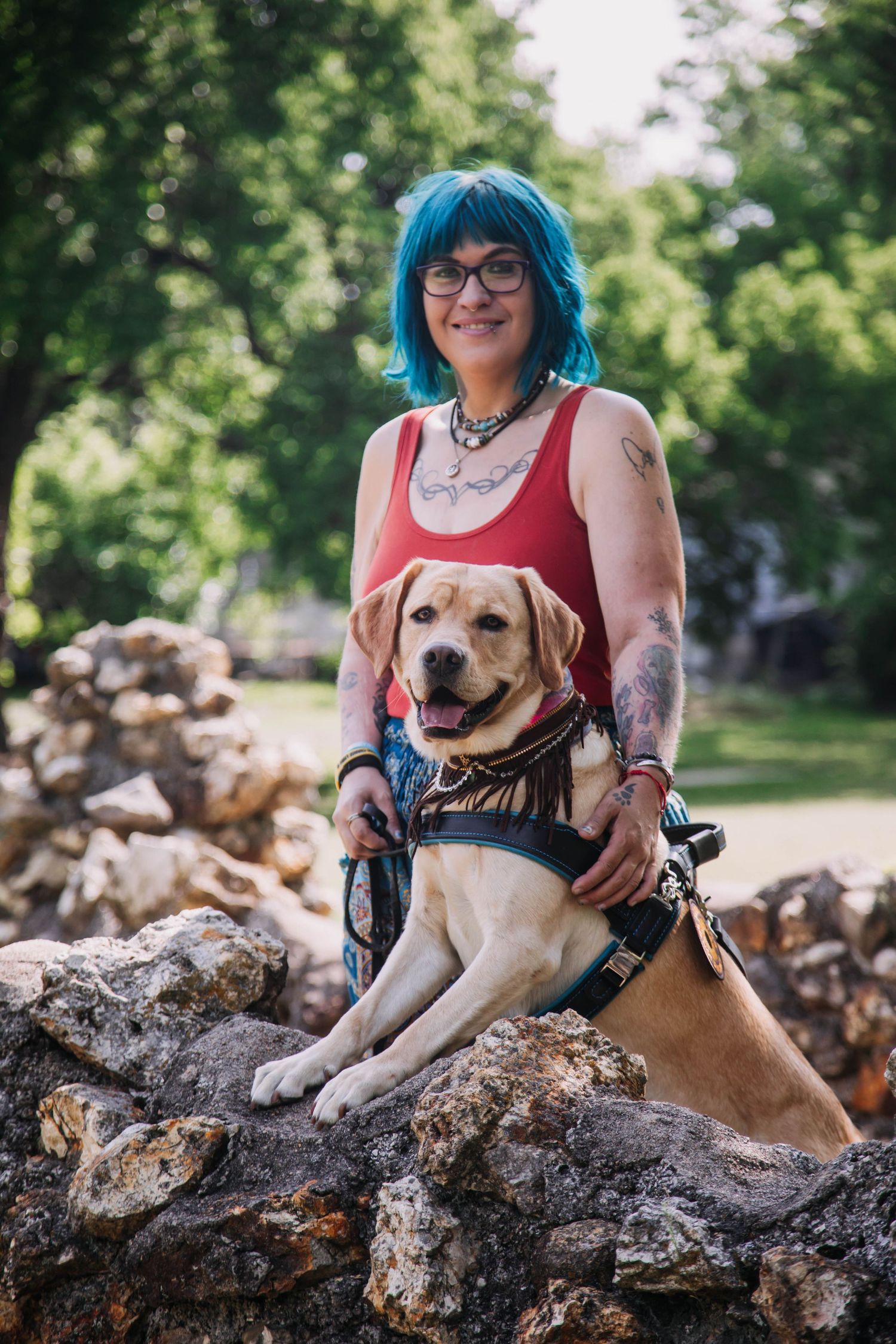Woman with blue hair and tattoos poses with yellow Labrador Retriever on rocky surface in sunny park.