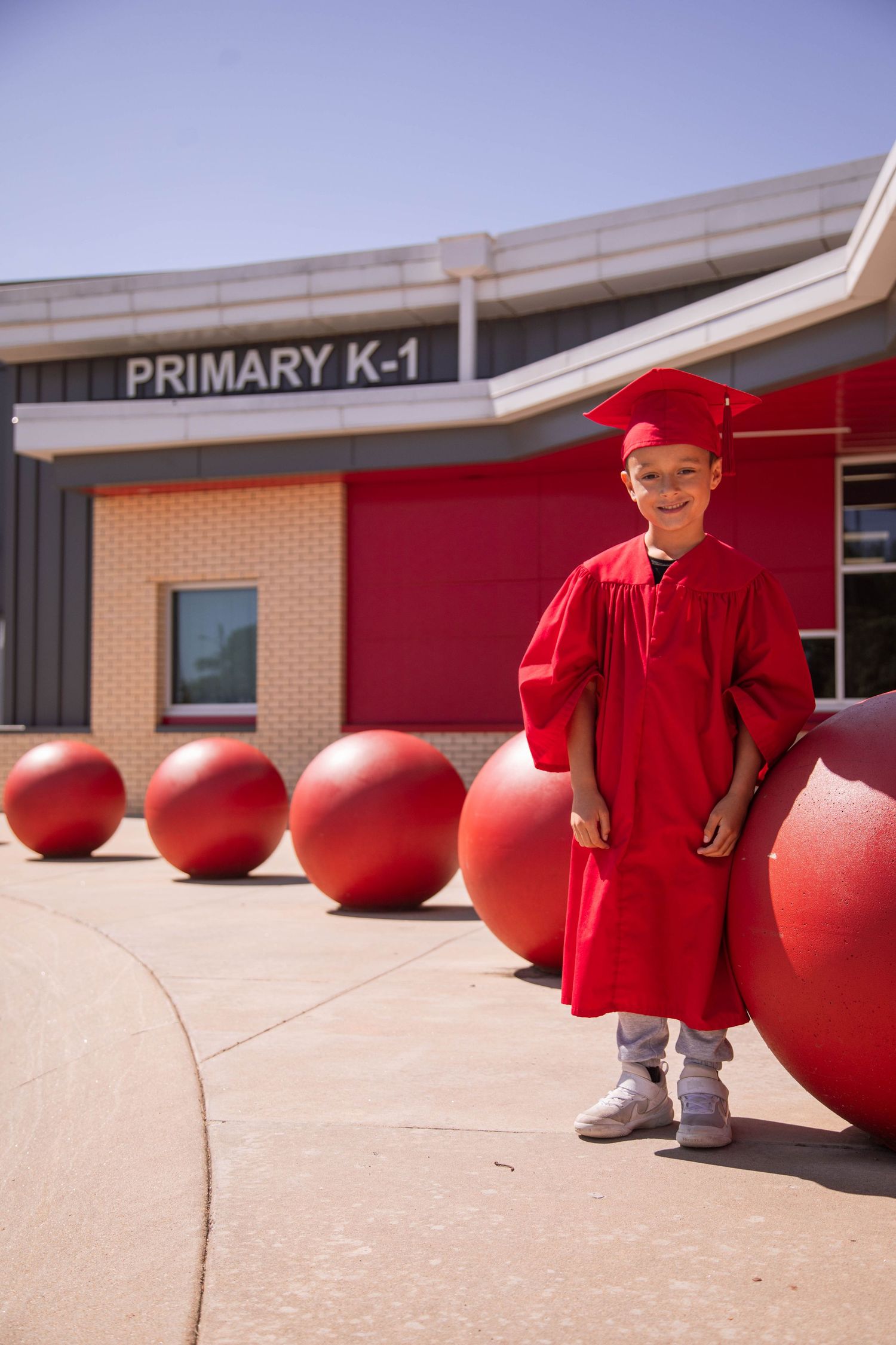 Young boy in red graduation cap and gown smiling outside Primary K-1 school building with red decorative spheres.