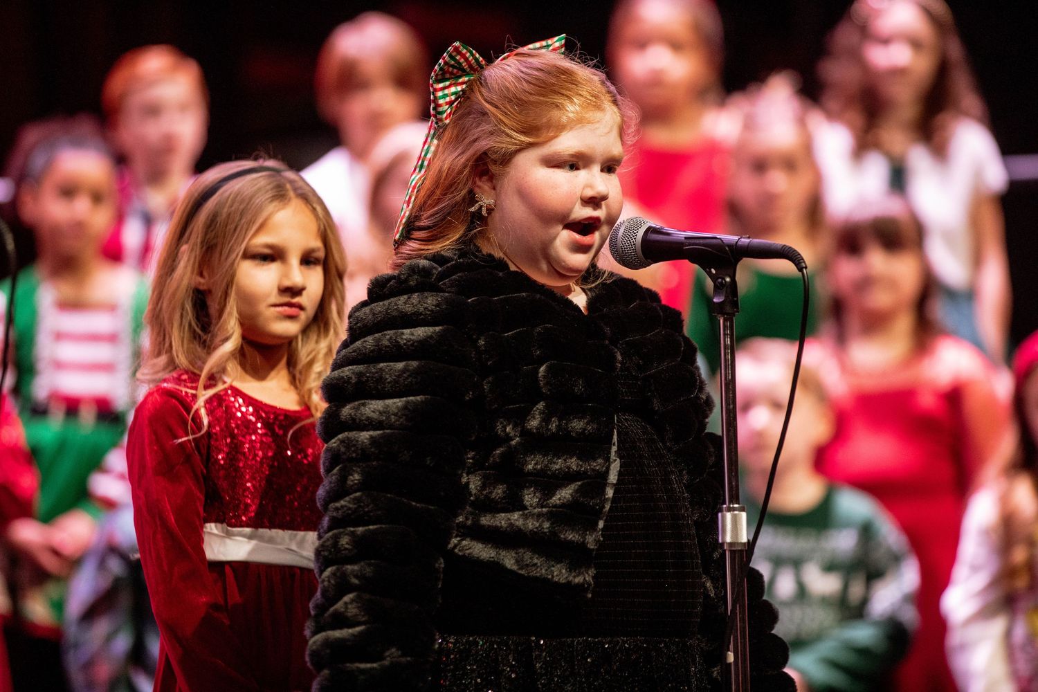 Young girl singing into microphone at Christmas choir concert, wearing black fur vest, with children in holiday attire behind her.
