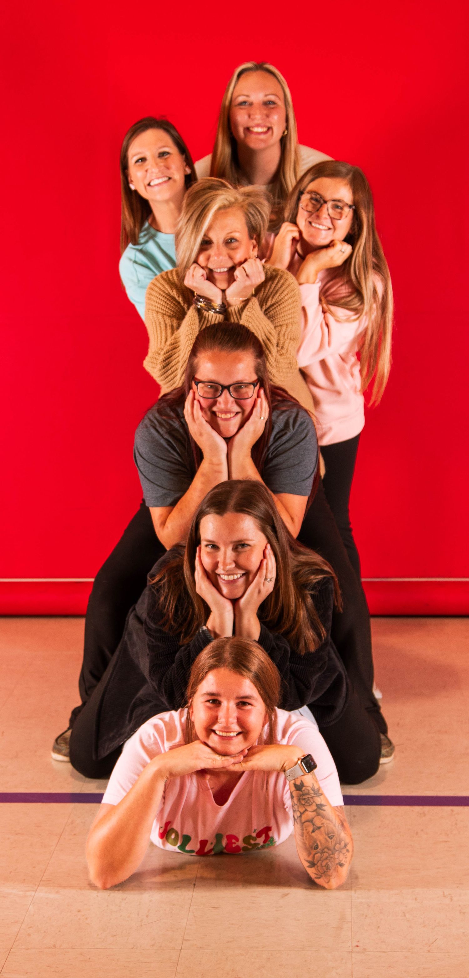 A group of seven women stacked in a cheerful pyramid pose against a red background.