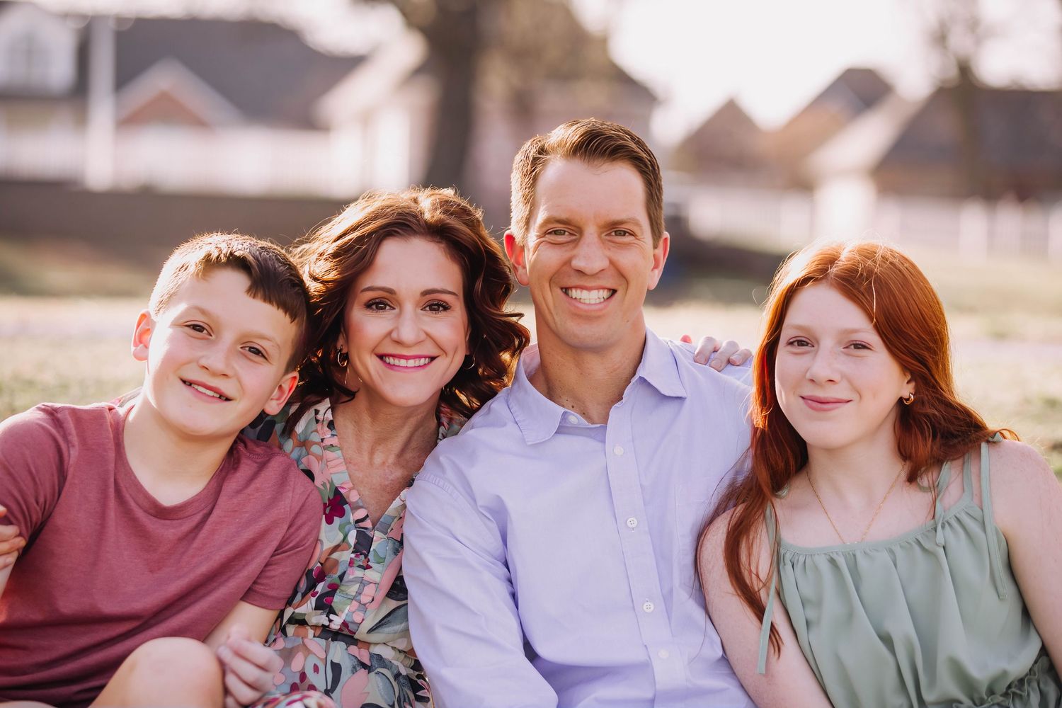 A smiling family of four poses outdoors at sunset, including two red-haired children and their parents.