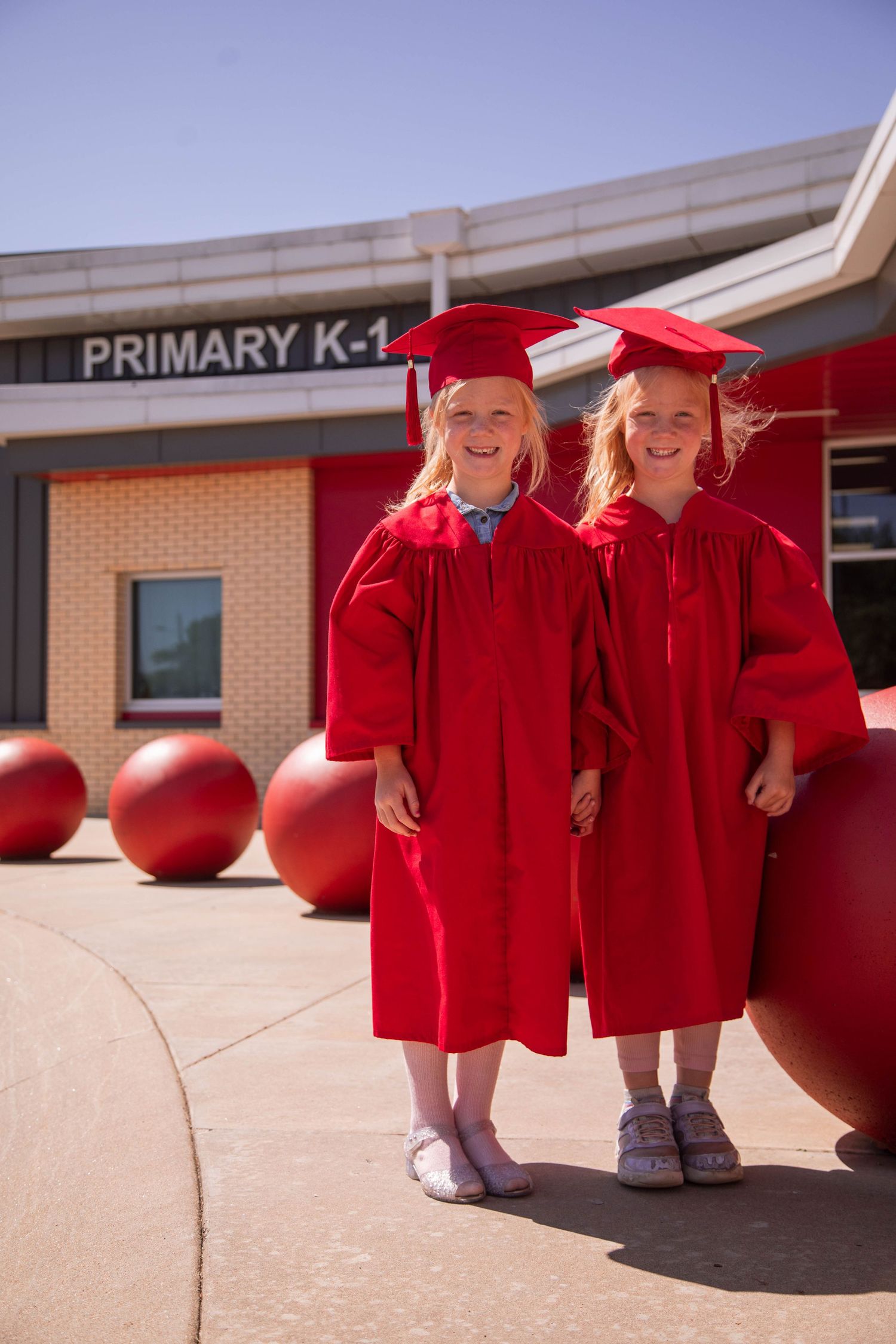 Two young girls in red graduation caps and gowns smiling outside a Primary K-1 school building.