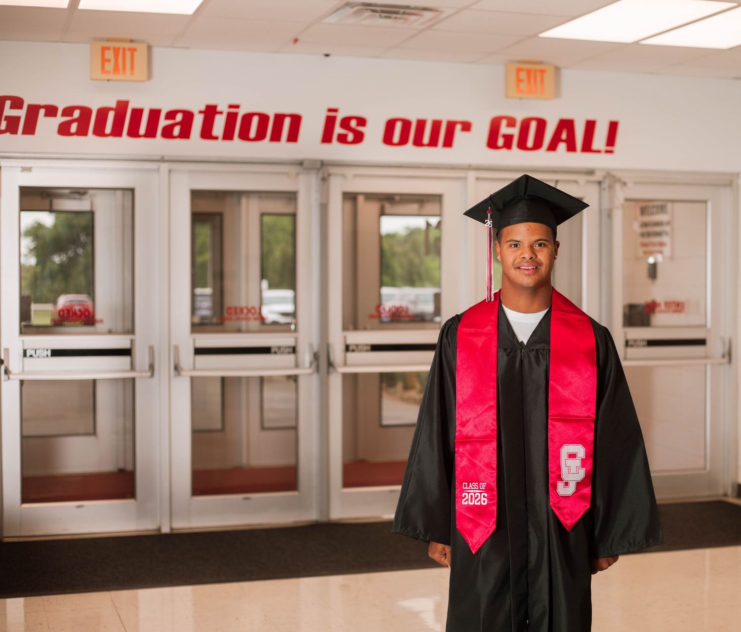 Graduate in cap and gown stands before school entrance with Graduation is our GOAL sign.