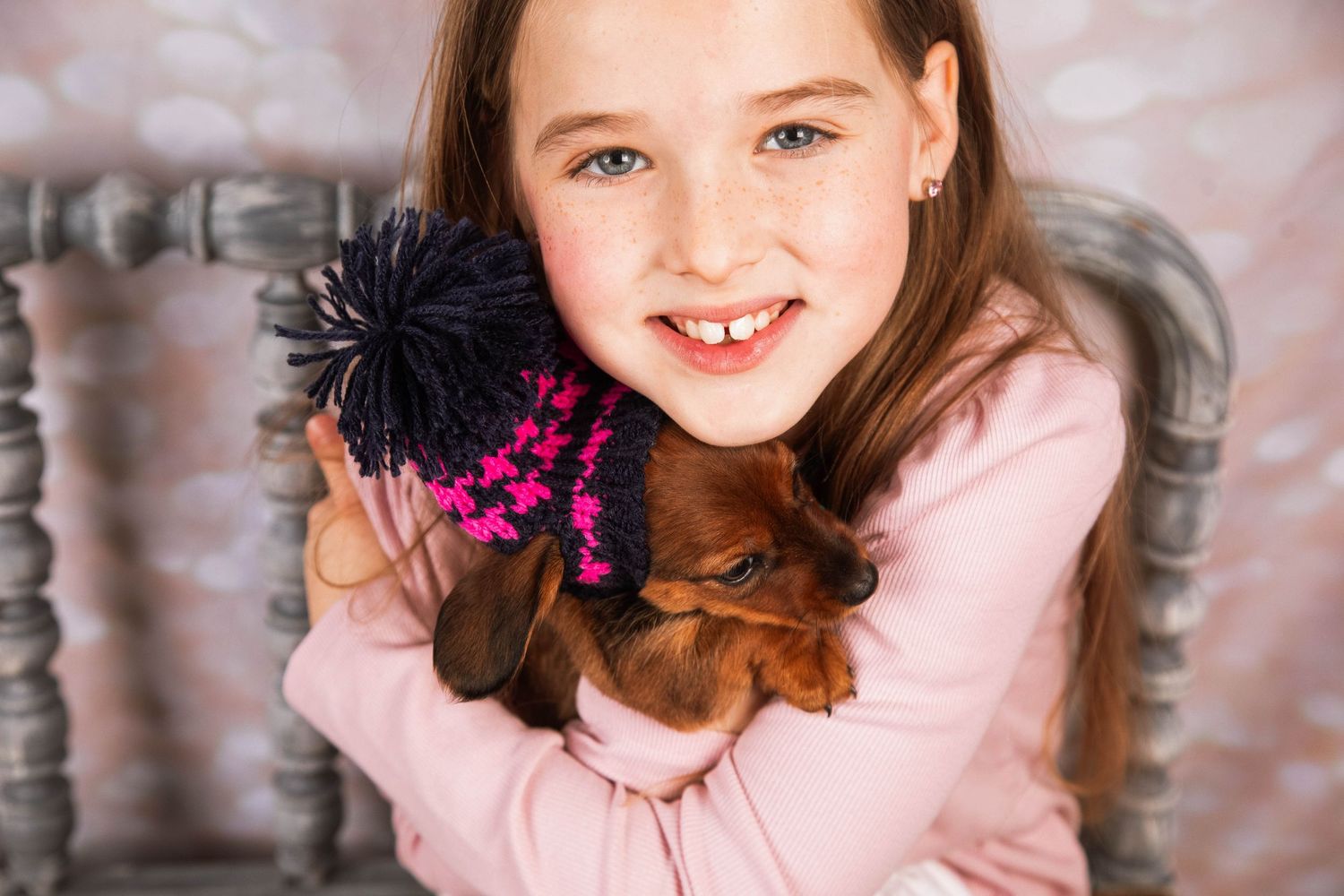 Smiling girl with freckles hugging a dachshund puppy, wearing a pink top with a decorative hair accessory.