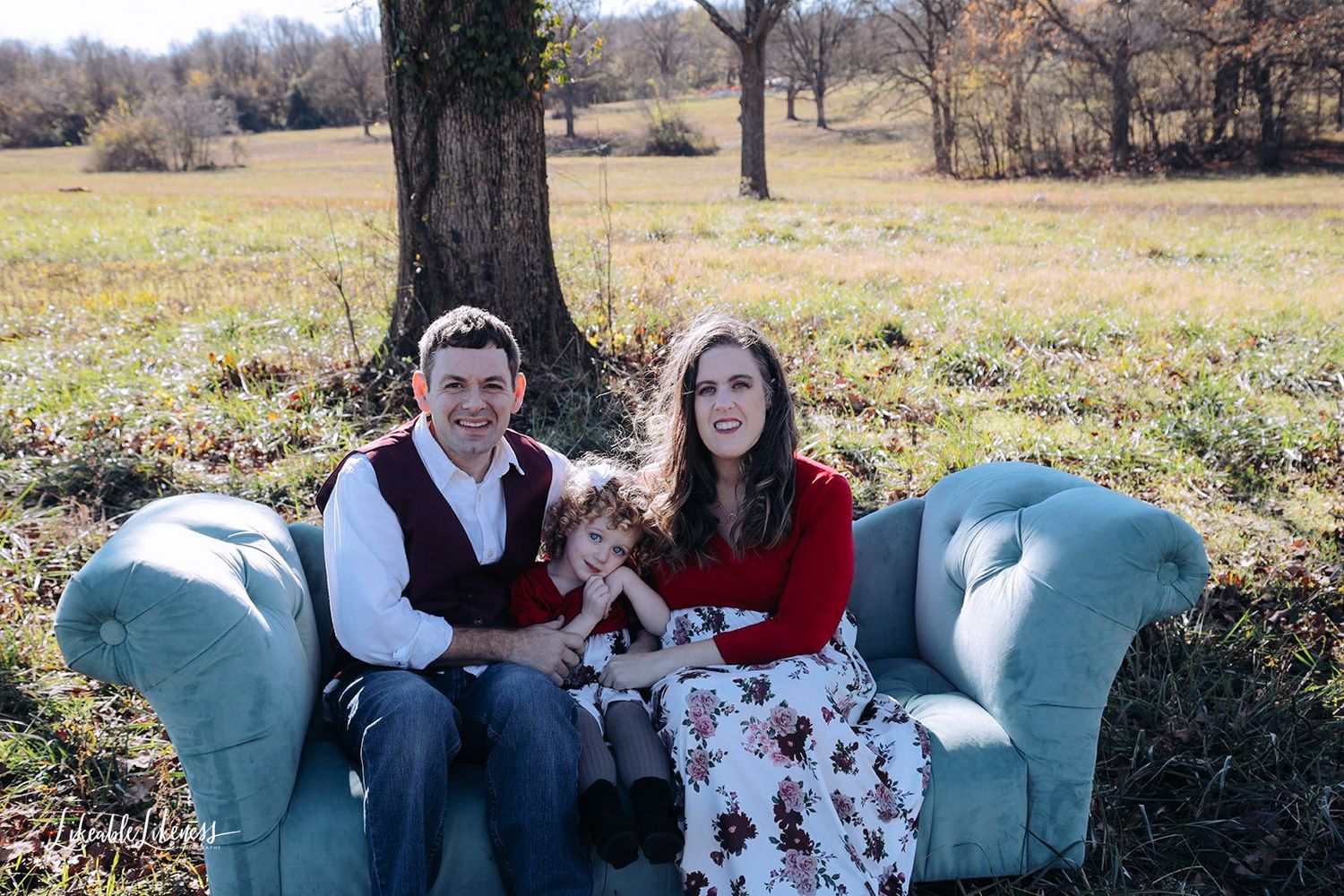 Young family of three poses on vintage teal sofa in rustic outdoor field during autumn.