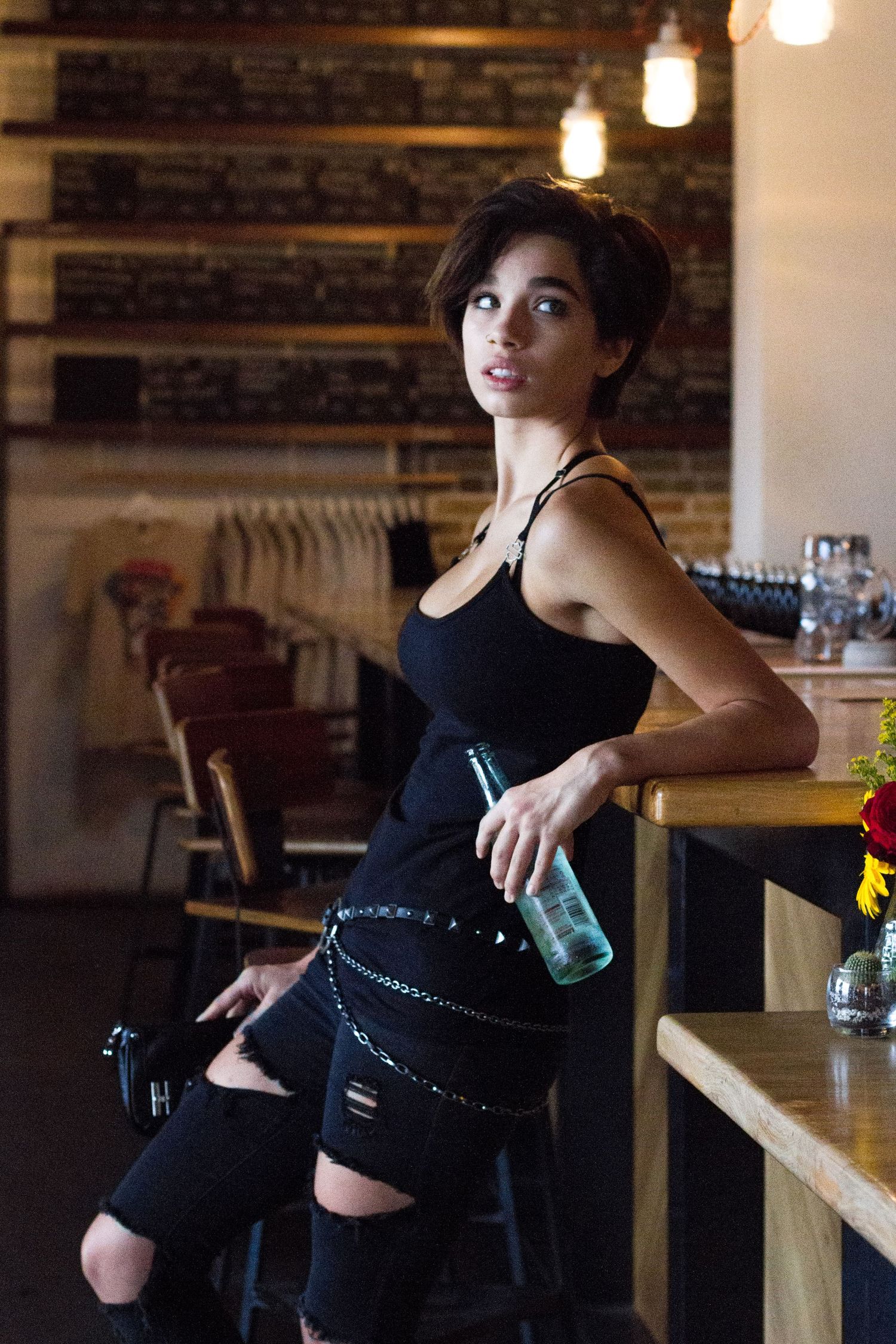 Someone in black attire posing against a wooden bar counter in a dimly lit restaurant setting.