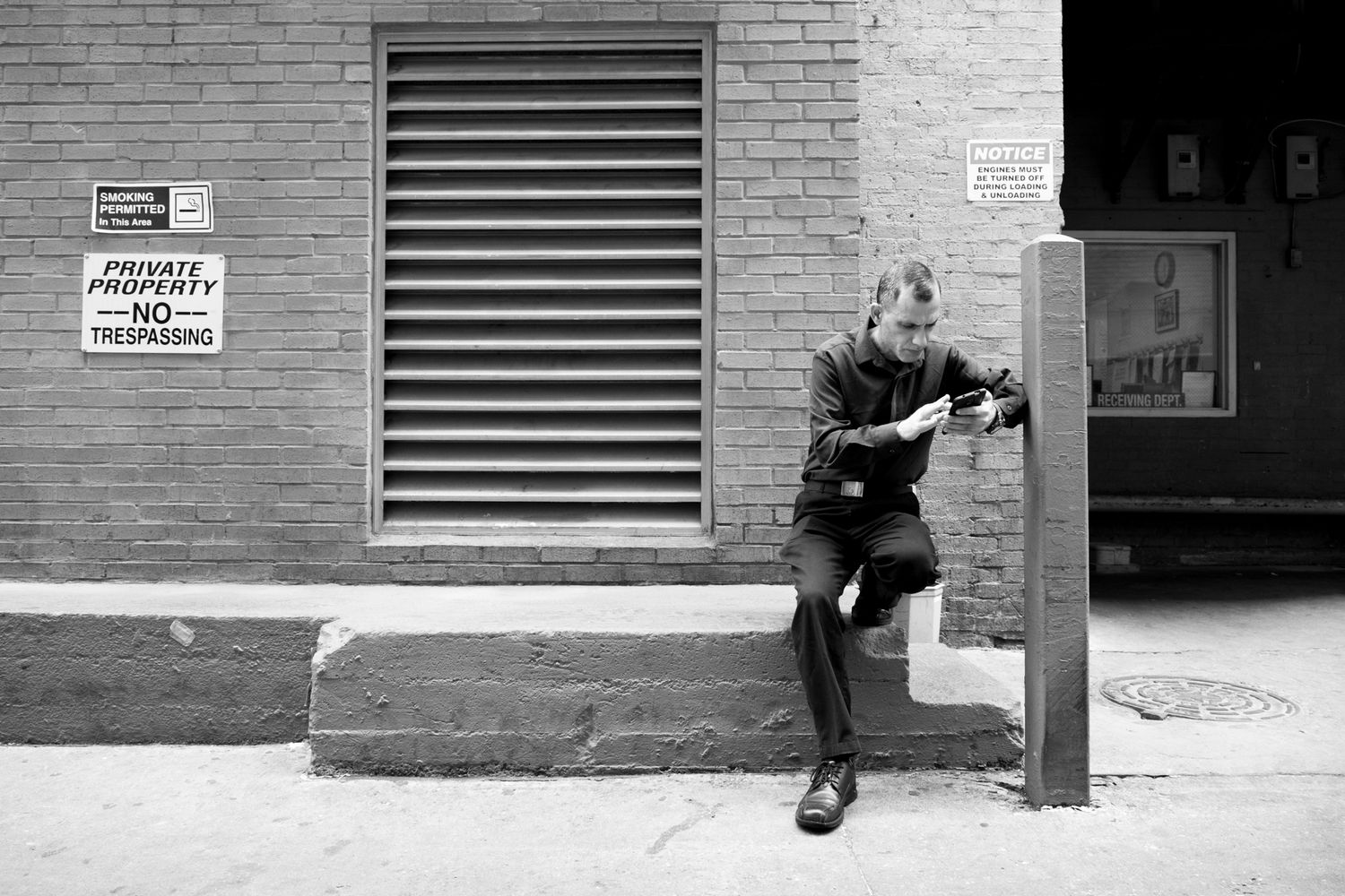 Black and white photo showing a figure sitting and reading near a brick wall with ventilation grates.