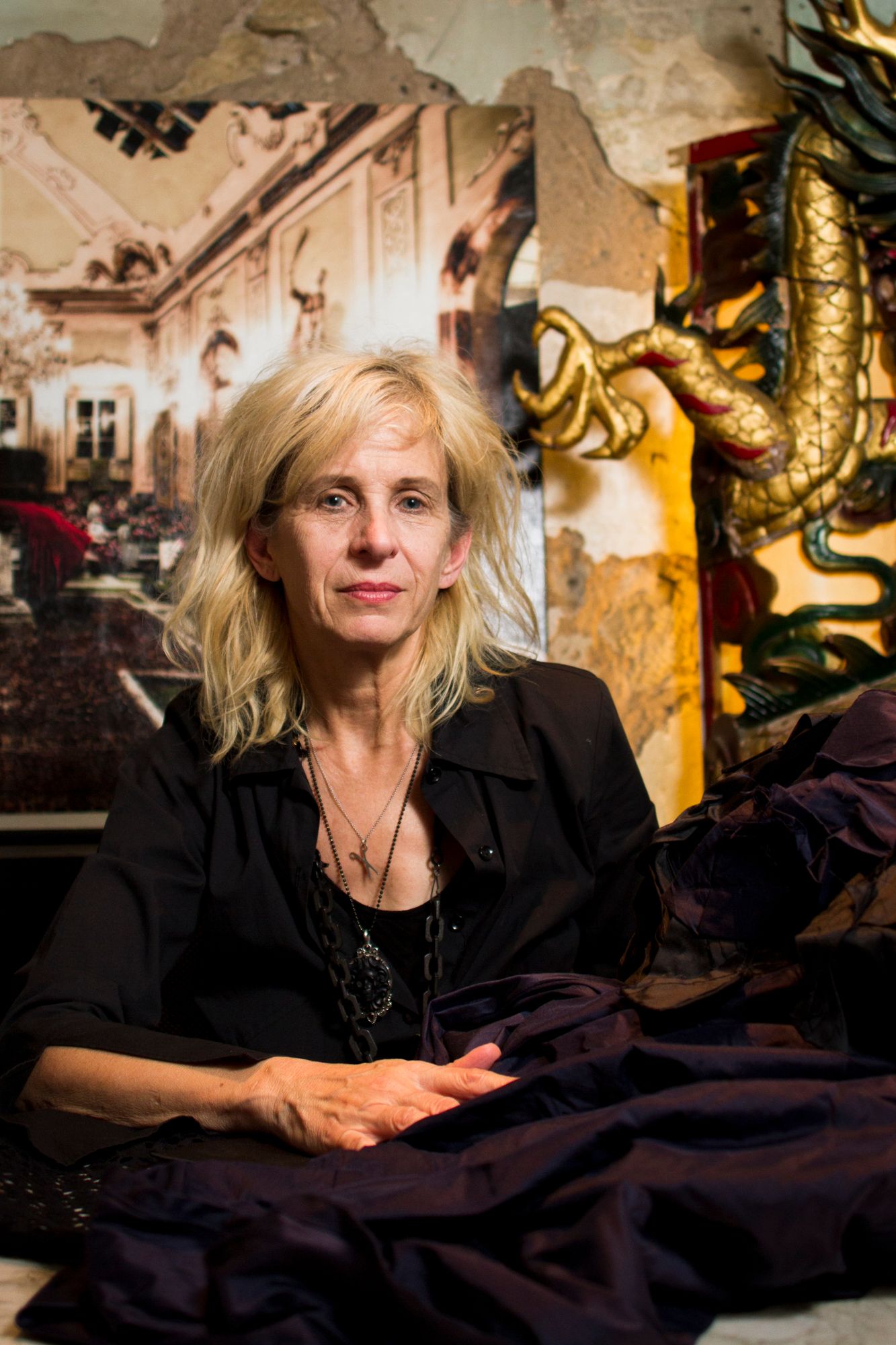 Designer, Gail Chovan, sits in a dimly lit room with ornate decorative elements and dark fabric visible.