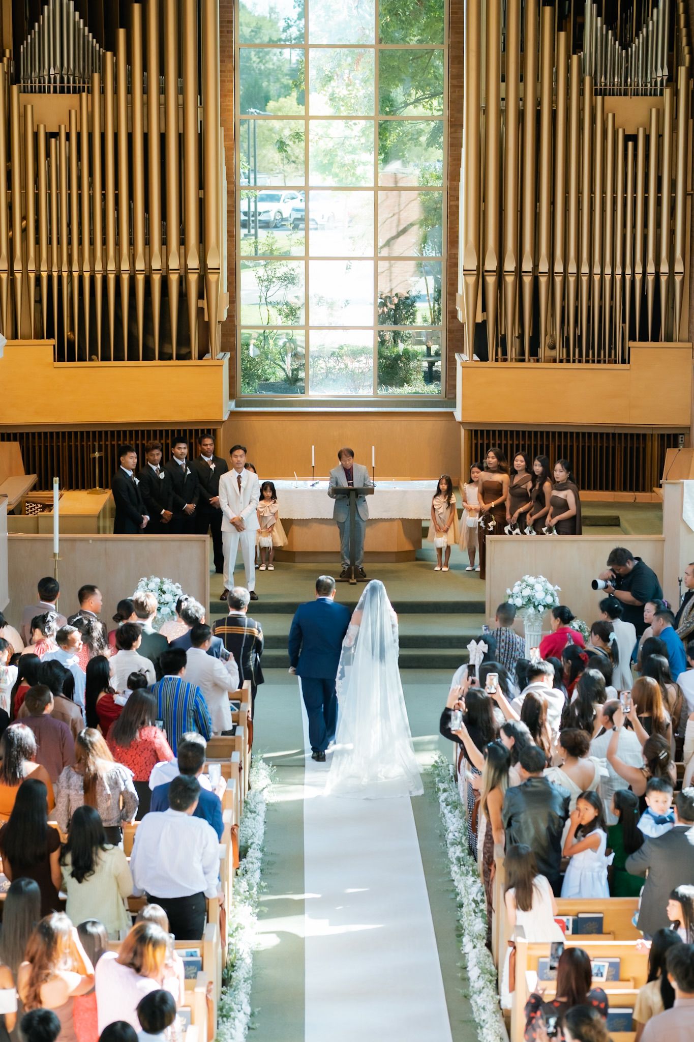 Bride walks down the aisle in a church wedding ceremony with guests seated in wooden pews and organ pipes behind altar.