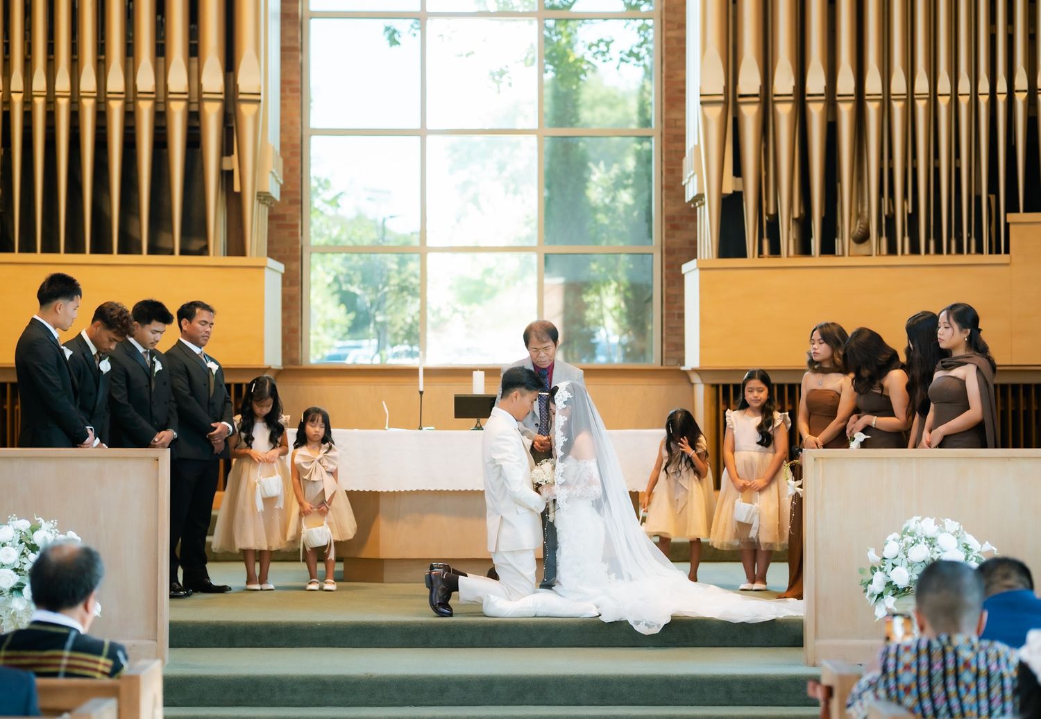 Bride and groom kneeling at church altar during wedding ceremony with flower girls and groomsmen in elegant chapel.