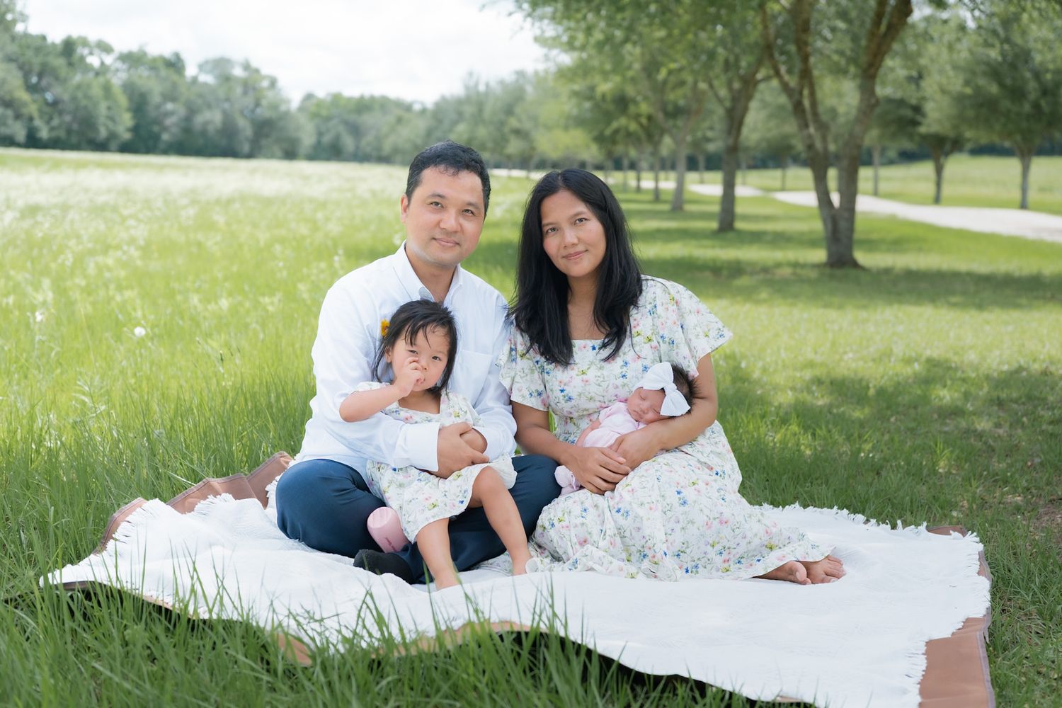 Family of four poses on white blanket in lush green park, parents smiling with toddler and newborn baby.