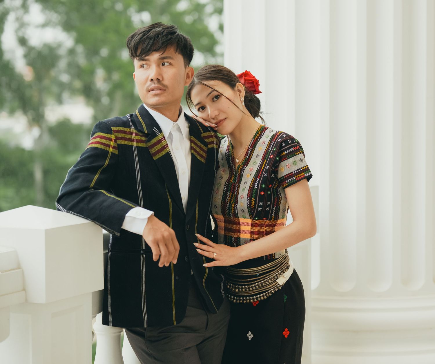 Couple in Hmong traditional dress share romantic moment by white columns.