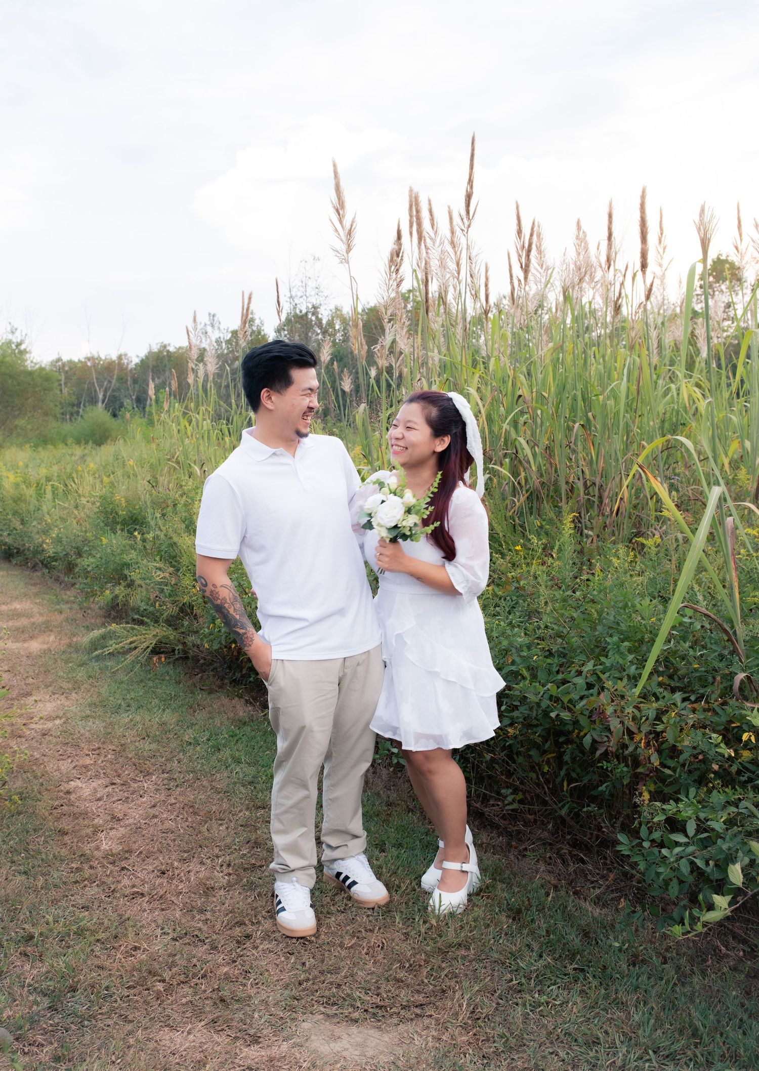 A couple laughs together while walking on a nature trail holding white flowers.