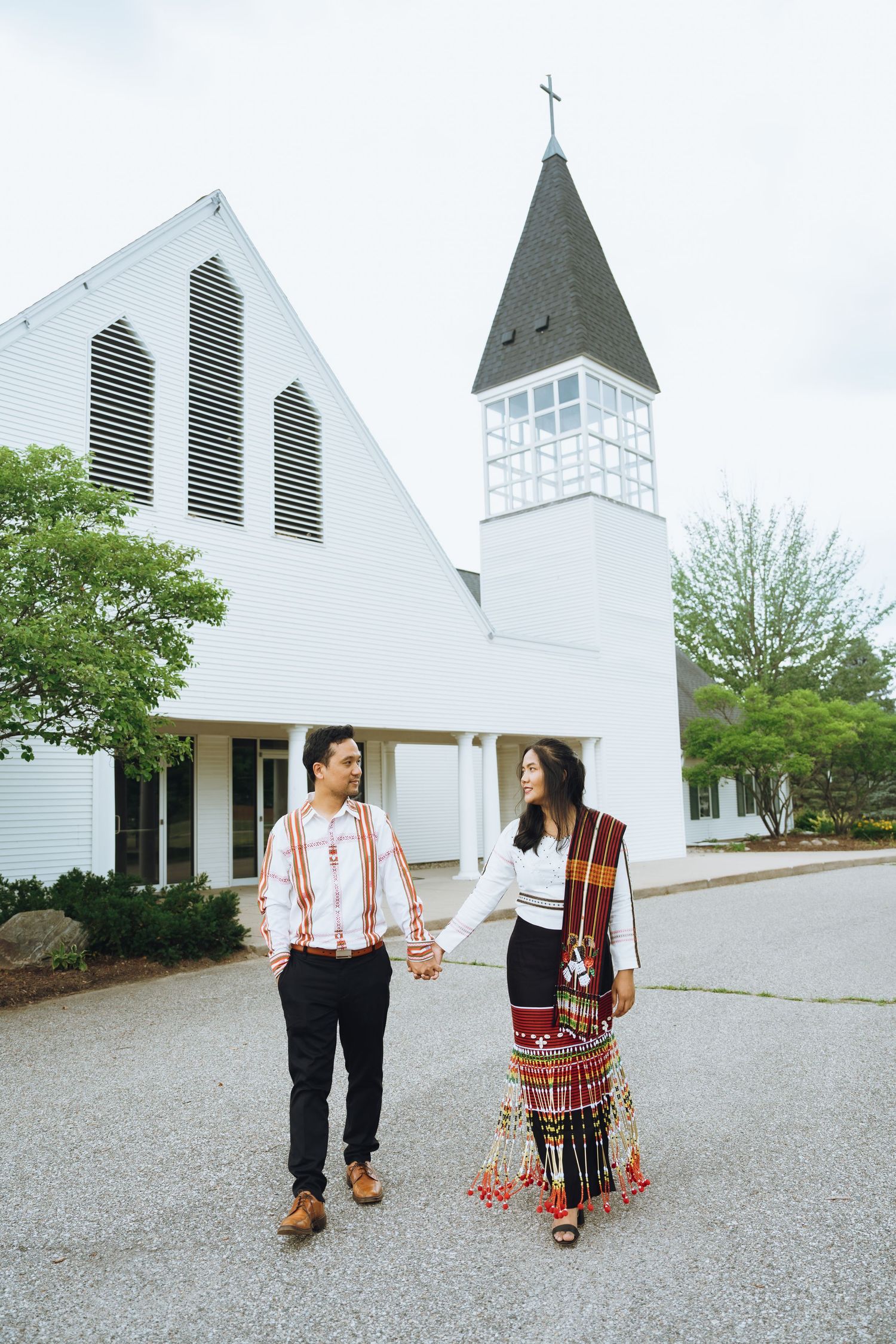 Couple in traditional attire walks hand in hand outside white church buildings.