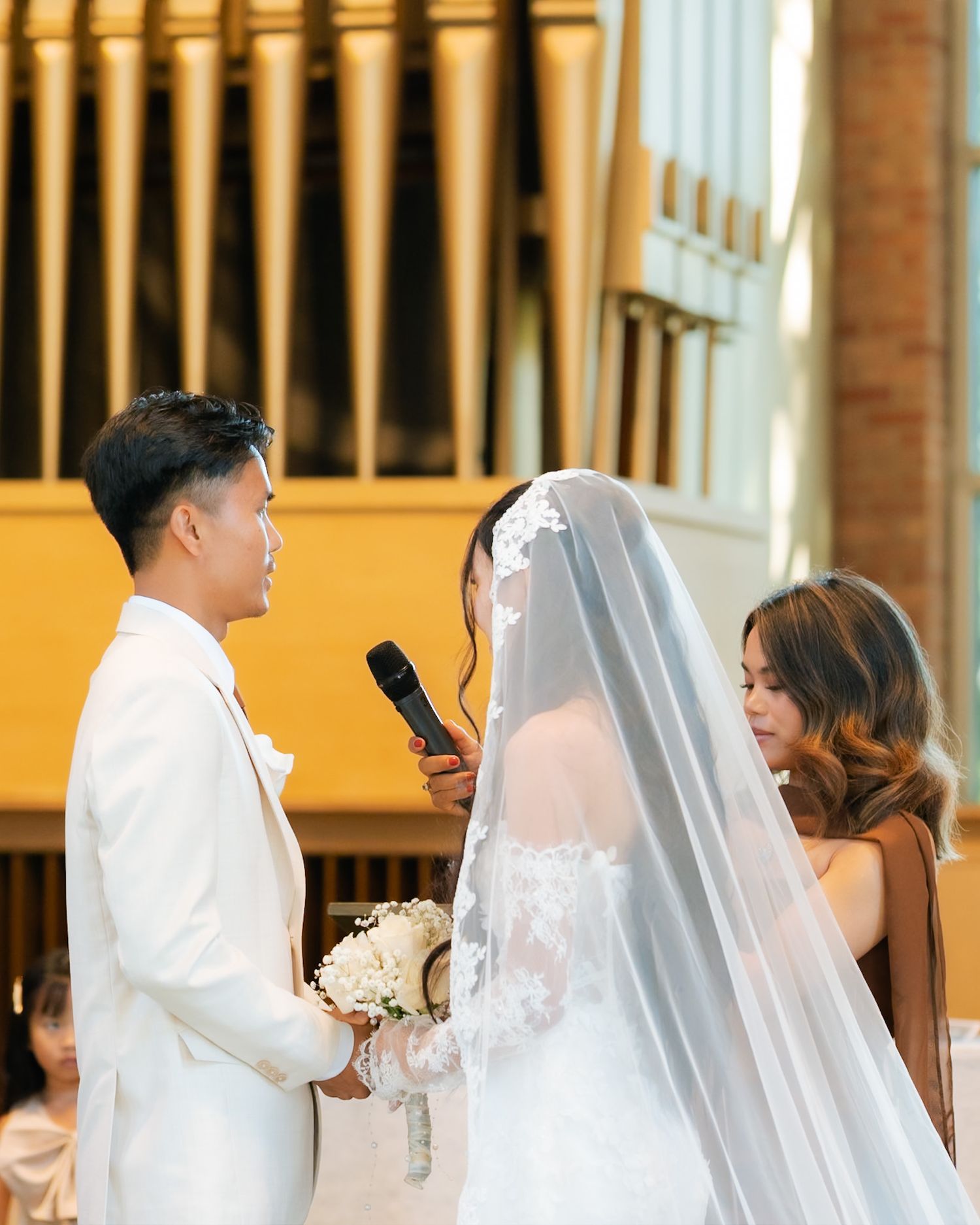 Bride and groom exchanging vows at church altar, bride in lace wedding dress with veil, groom in white suit.