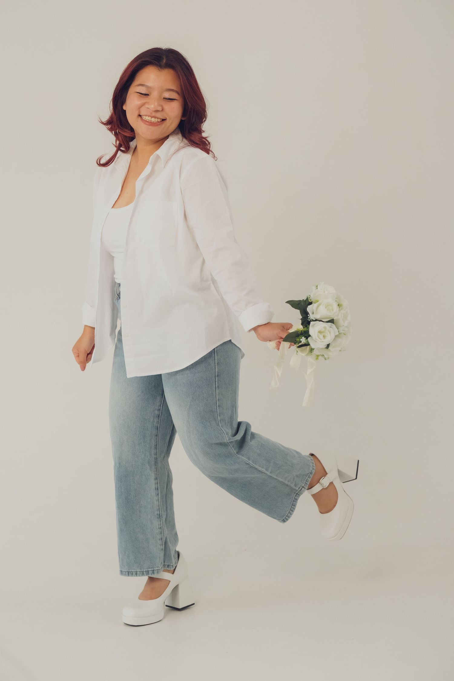 Woman in white button-down and jeans dances joyfully with flowers against neutral backdrop.