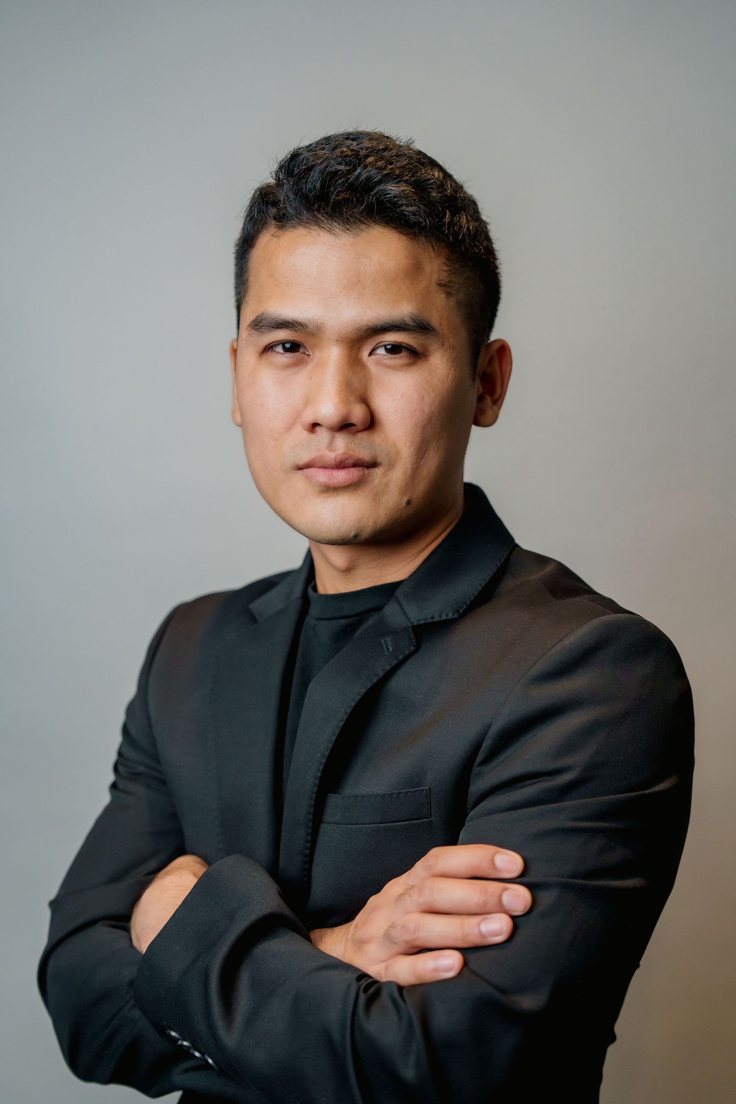 Professional headshot of person in black blazer with arms crossed against neutral background.