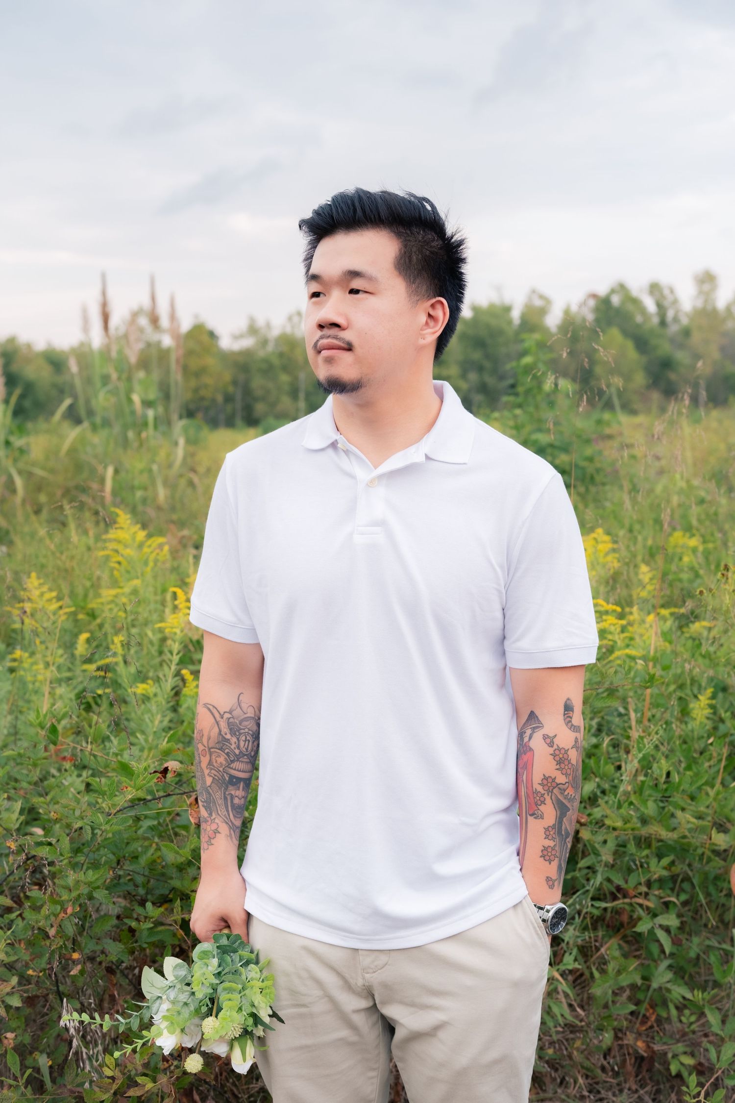 A person in a white polo shirt with tattoos poses in a field of wildflowers.