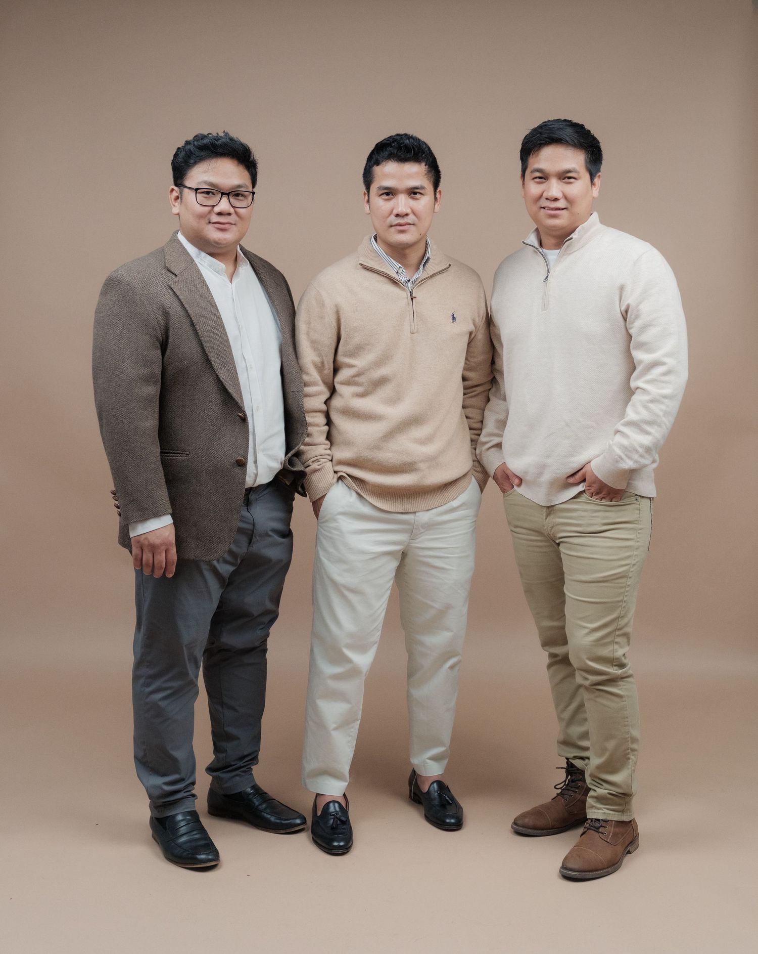 Three people in smart casual wear stand together against a brown background for a formal portrait.