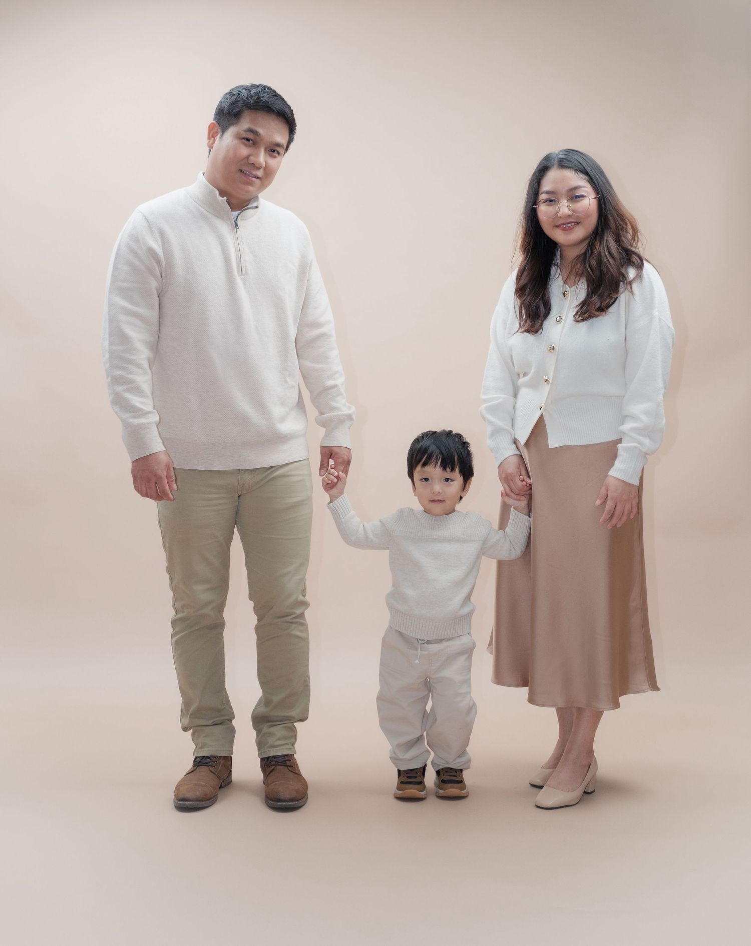 Family holds hands in coordinated cream outfits during studio photoshoot.