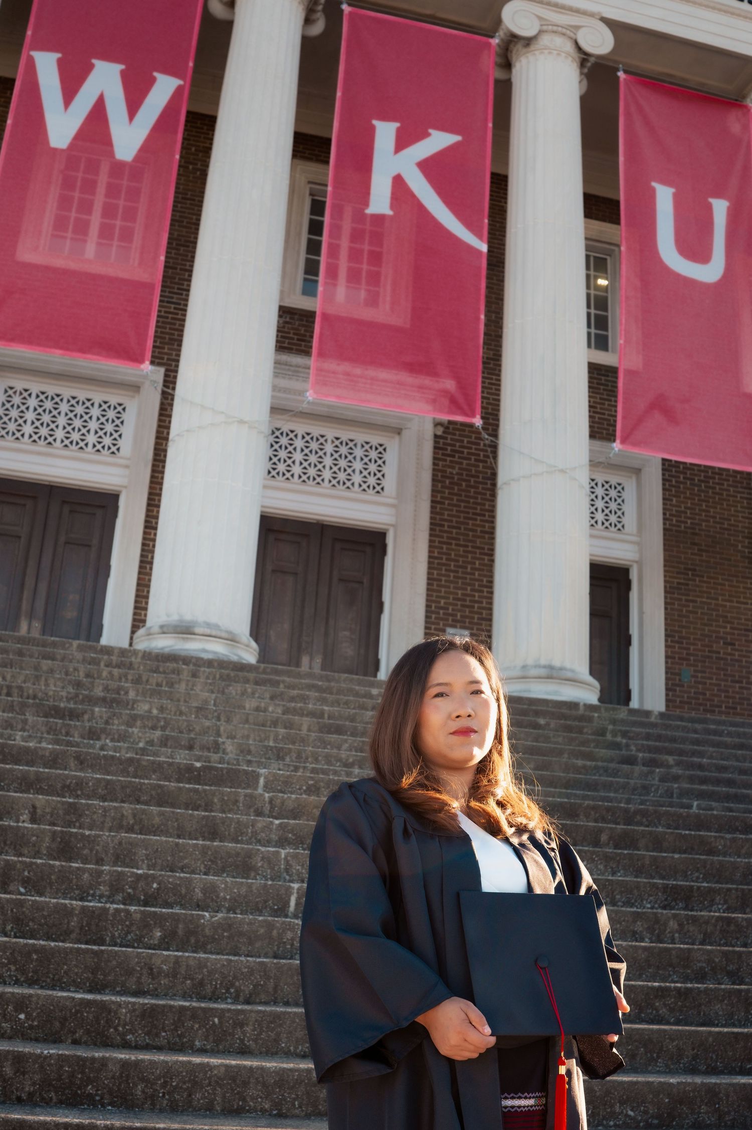 Graduate in cap and gown posing in front of red WKU banners on campus steps.
