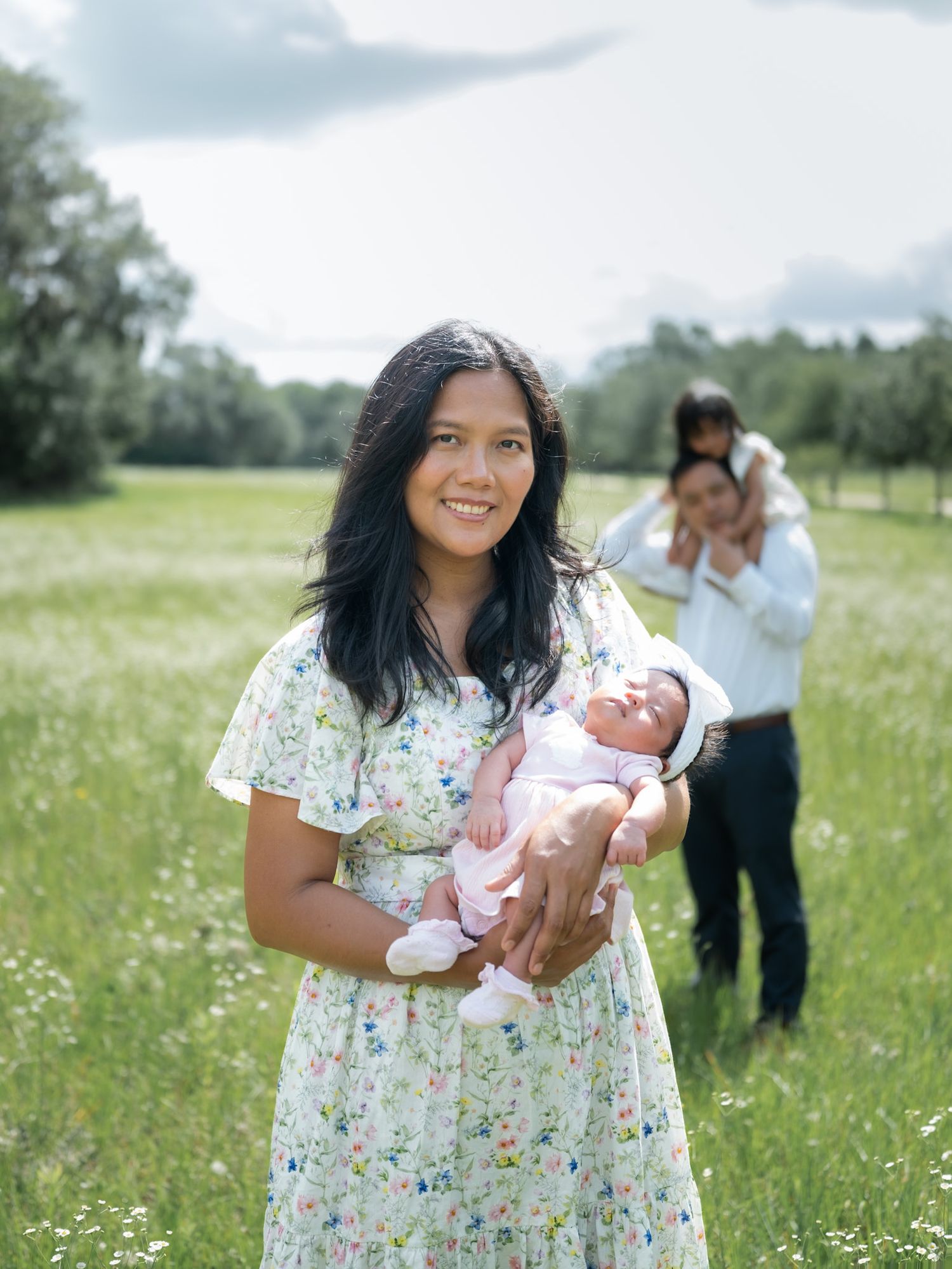 A smiling woman in a floral dress holds a newborn in a green meadow, with a man and child in the background.