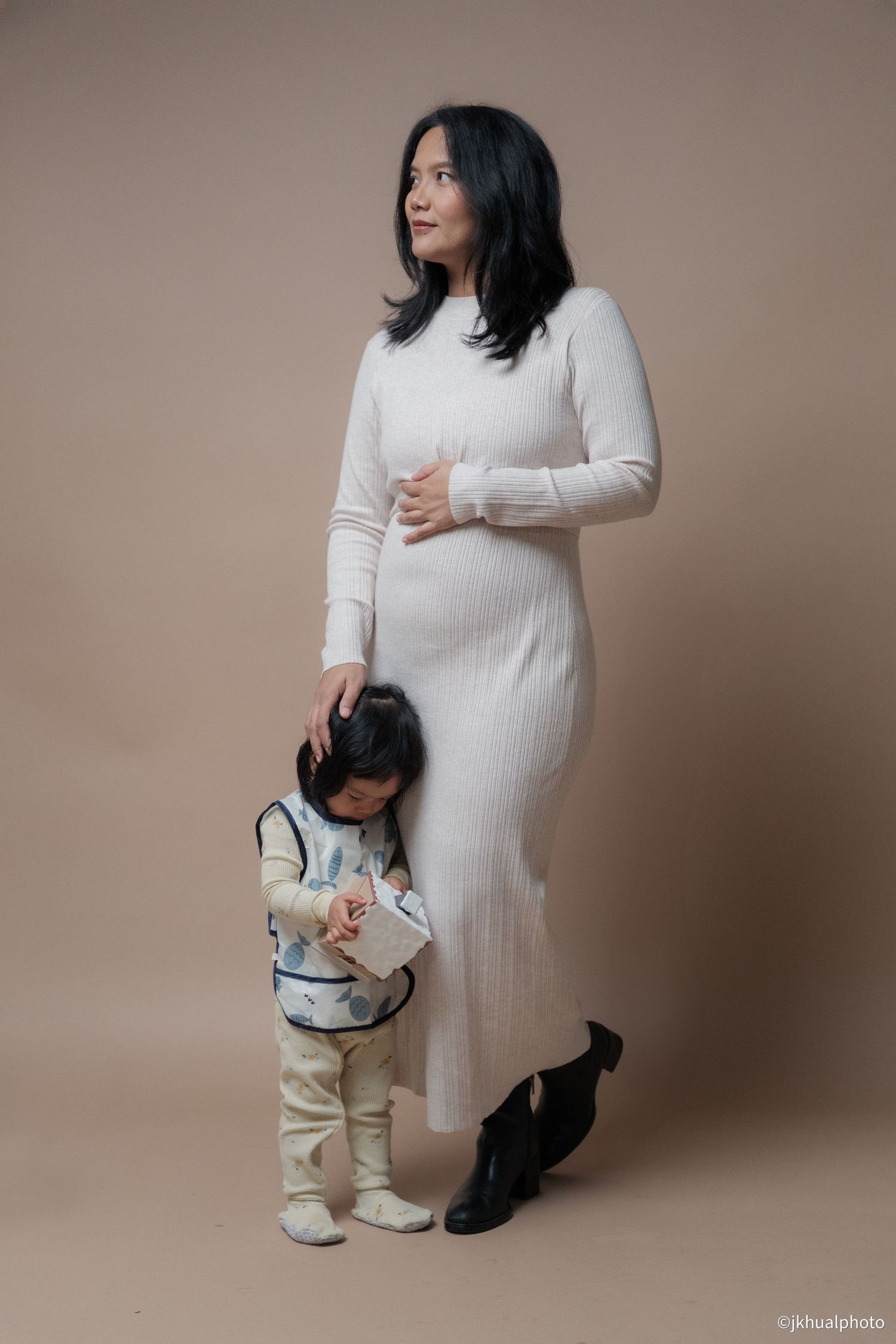 A maternity photo session featuring a woman in white with a small child in casual attire against a beige background.