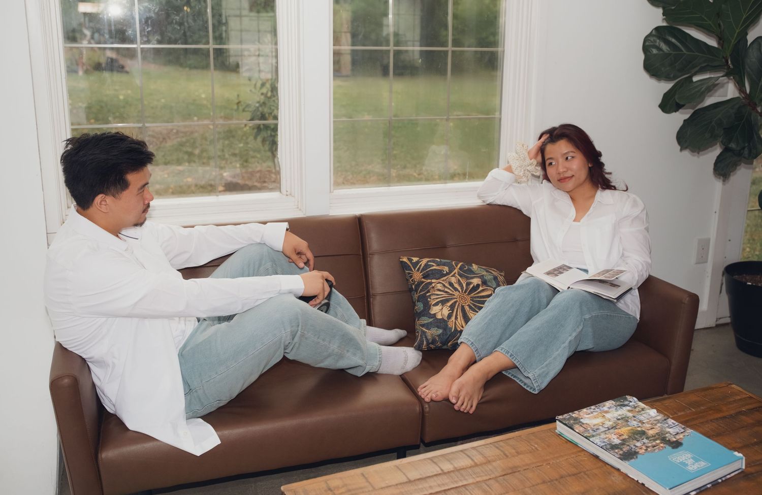 Two people in coordinated outfits relax and play on a brown leather couch by large windows.
