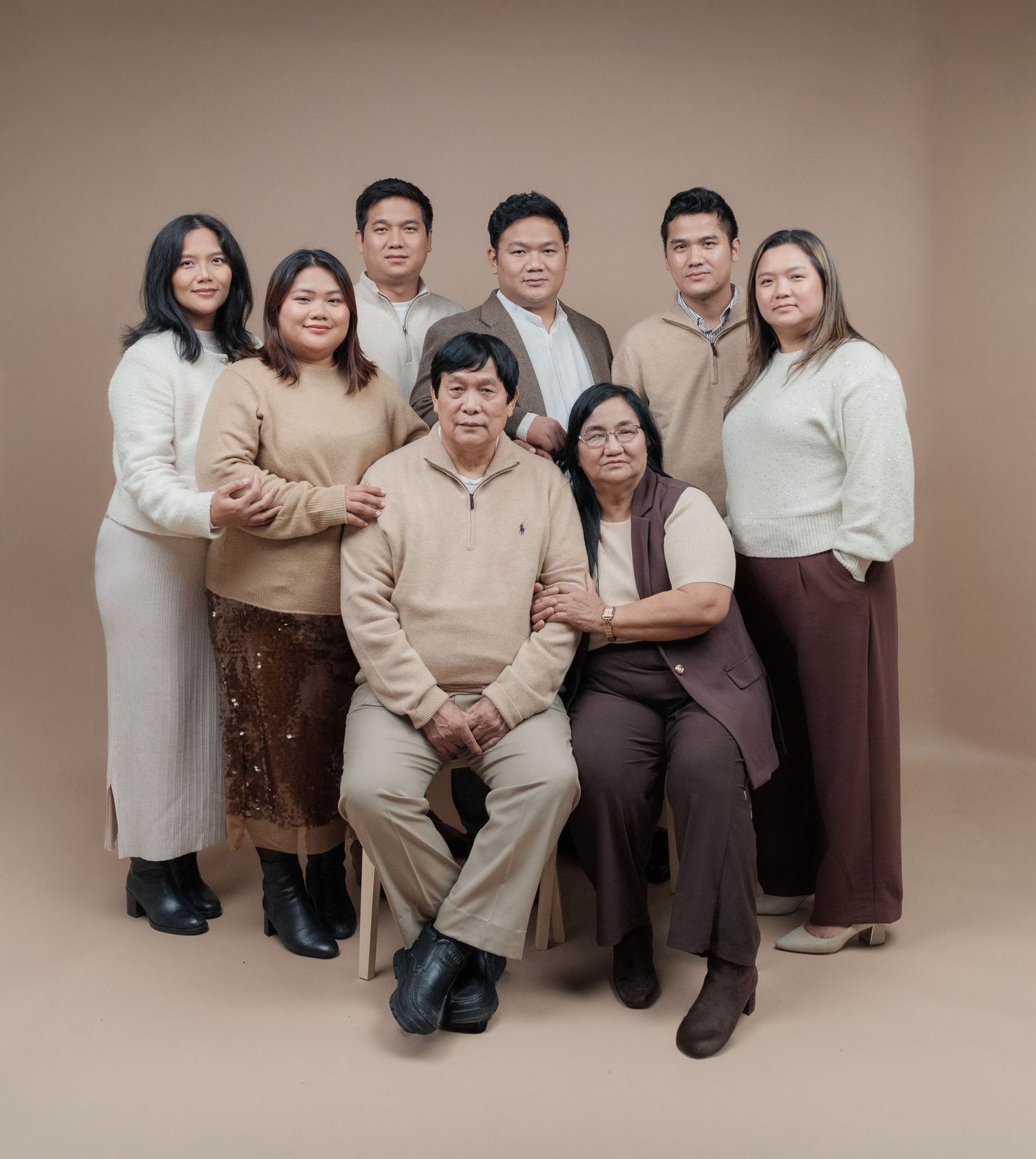 Studio portrait of family members in coordinated neutral tones sitting together on brown backdrop.