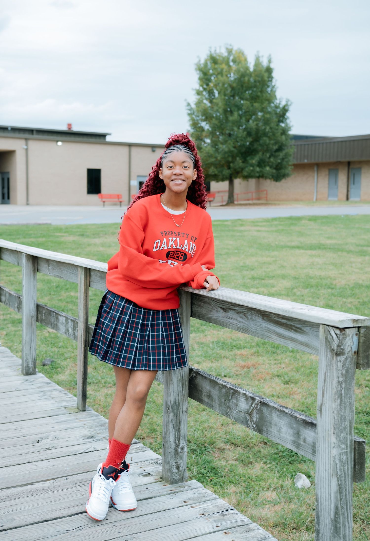 Student in red sweater and plaid skirt poses on wooden deck at school.