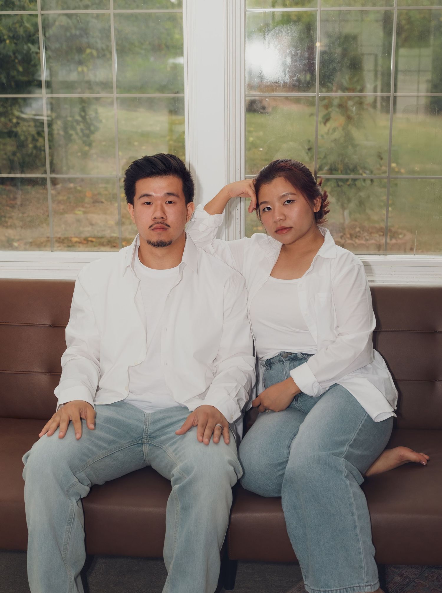 Couple in coordinated white shirts and jeans sits casually on brown leather sofa.
