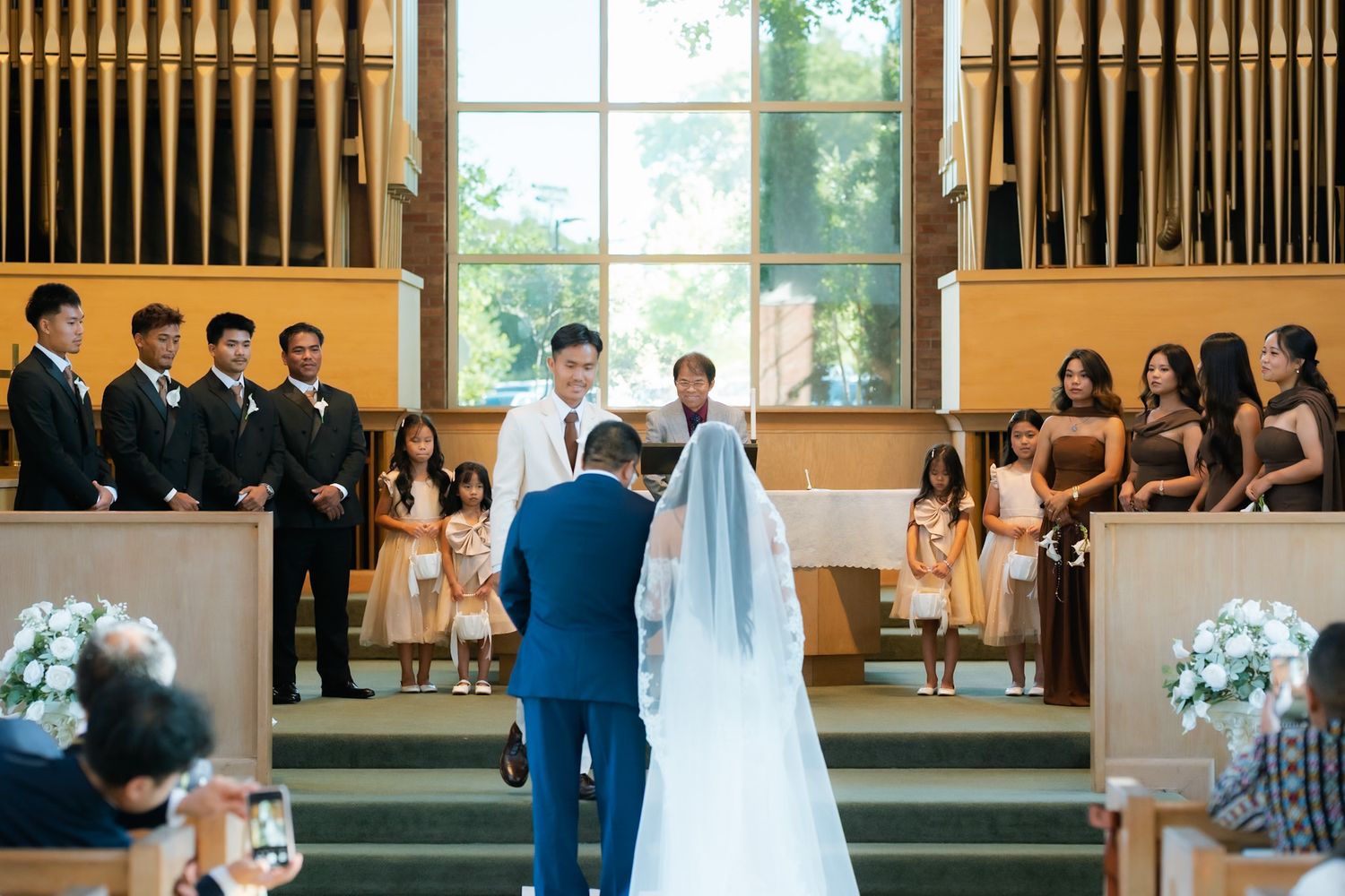 A bride and groom exchange vows at a church altar surrounded by groomsmen, bridesmaids, and flower girls during a wedding ceremony.